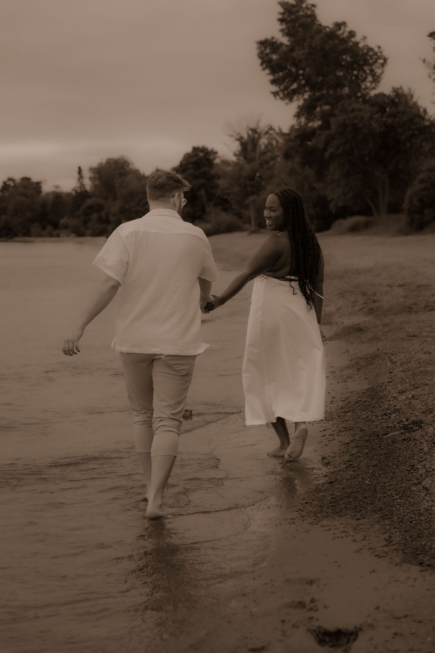 A couple walks hand in hand along the water's edge at dusk.