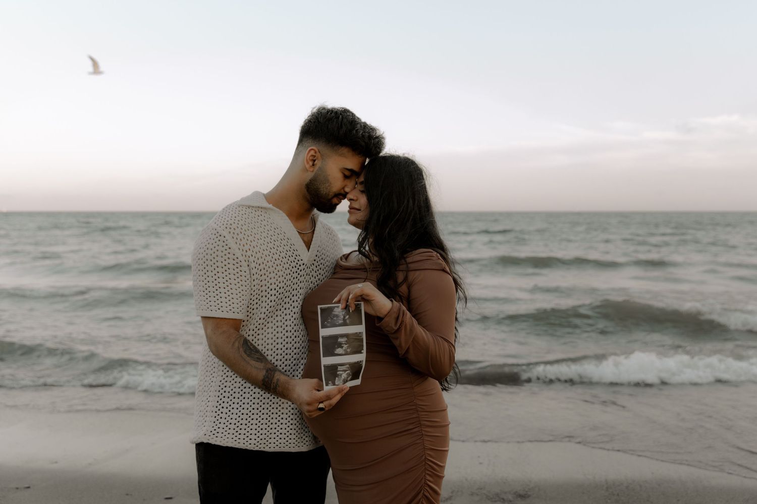 Couple sharing pregnancy announcement moment on beach with ultrasound photos at dusk.