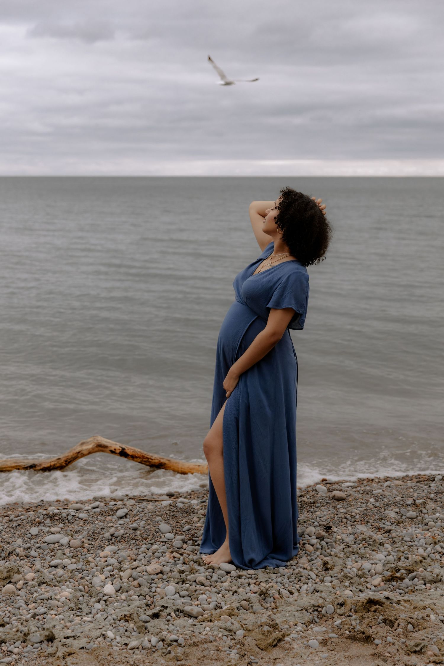 Pregnant woman in blue maxi dress stands on rocky beach gazing at calm sea under cloudy sky with seagull flying overhead.