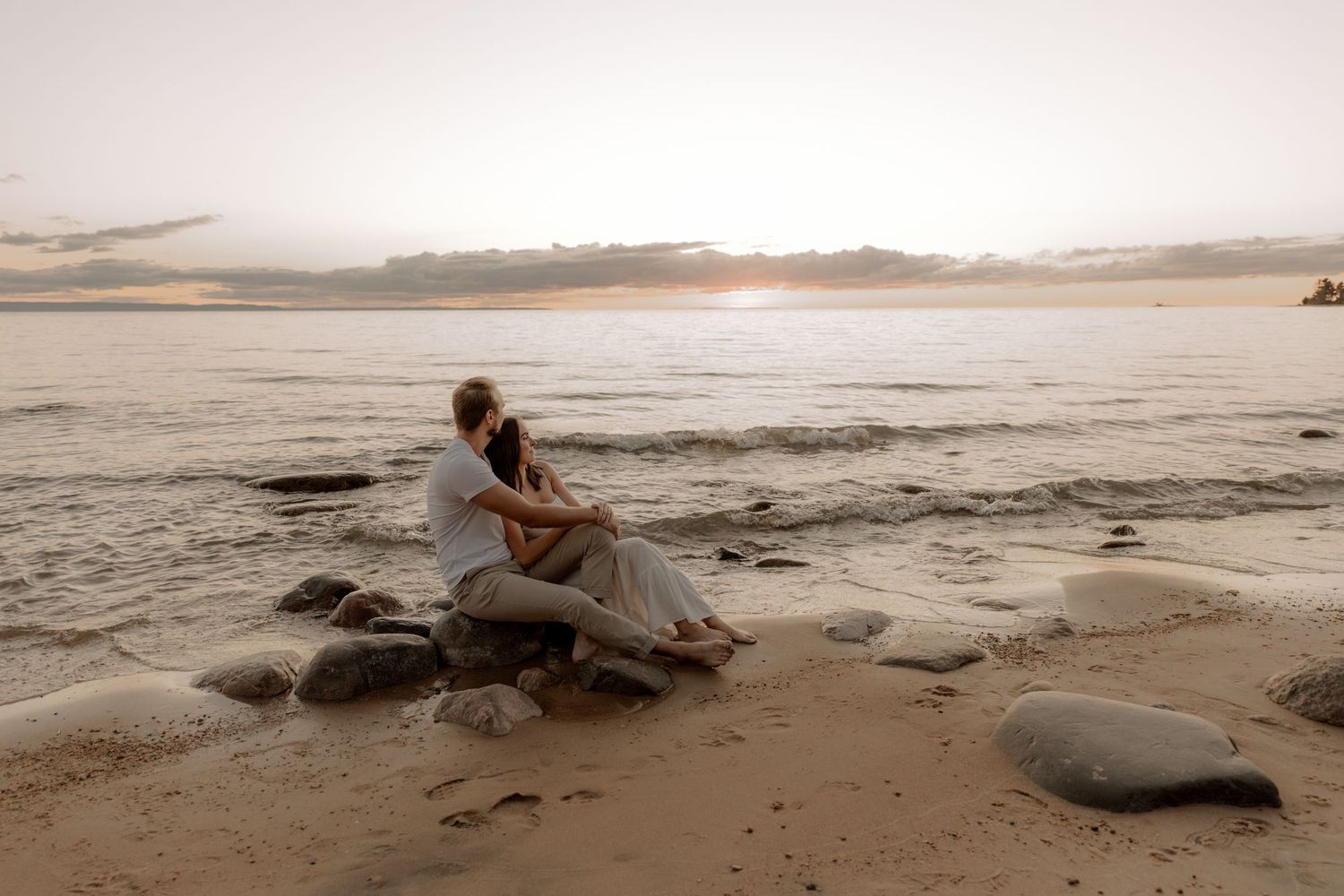 Couple sitting on rocks embracing at sunset on a serene sandy beach with calm ocean waves in the background.