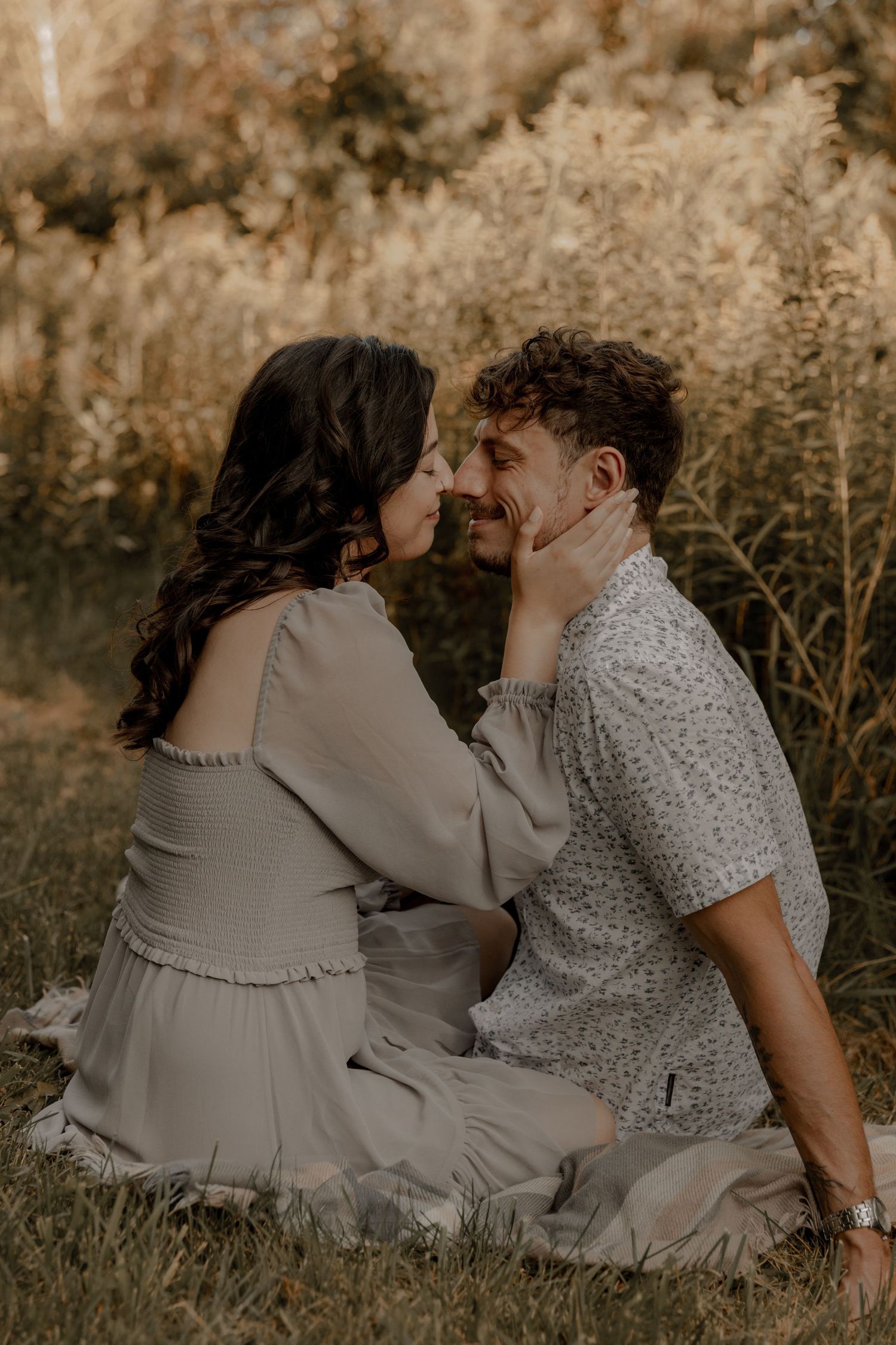 A couple sits in a golden field, sharing a tender moment as the woman gently cups the man's face.