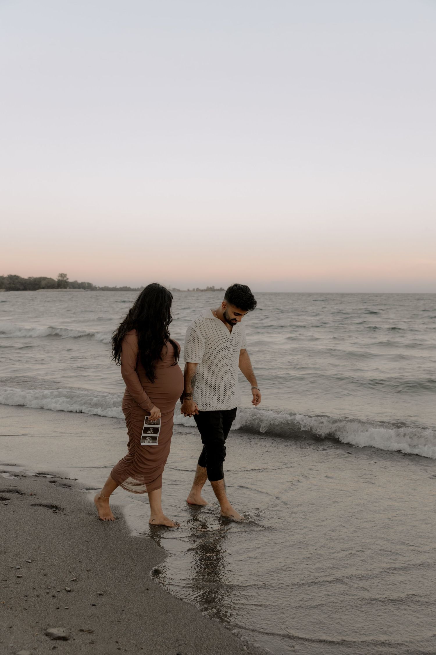 Couple wading in ocean waves while holding ultrasound photos during sunset.