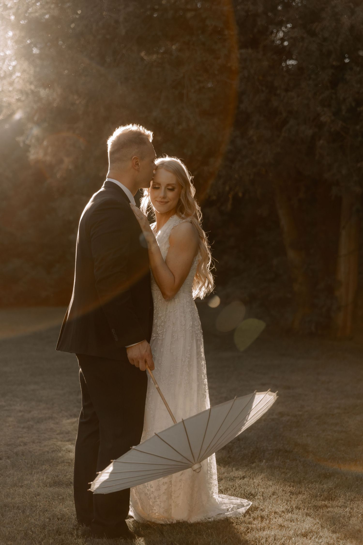 A romantic wedding couple embraces in golden sunlight with a white umbrella resting on the ground beside them.