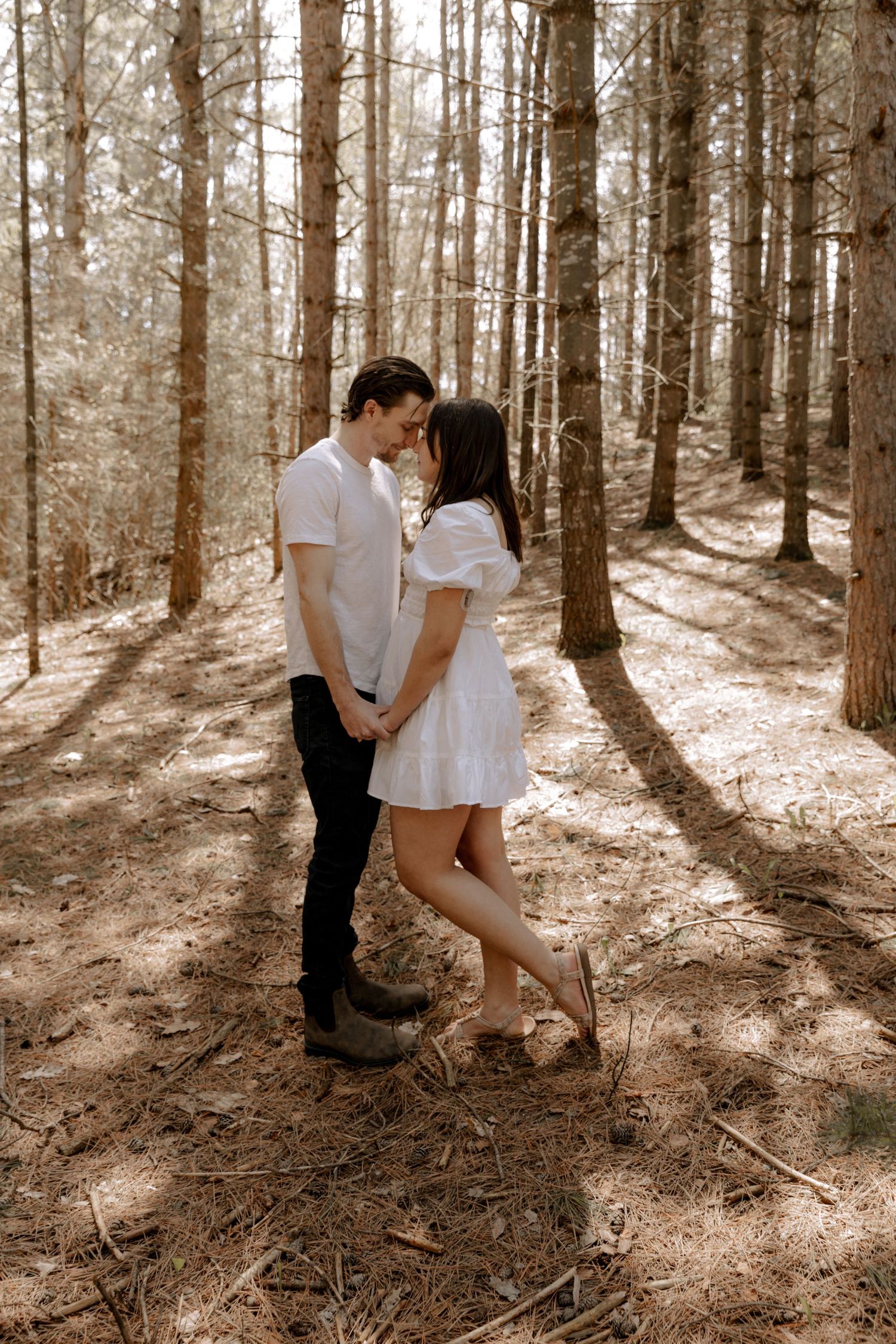 Romantic couple embraces in sunlit forest setting with white dress and casual attire.