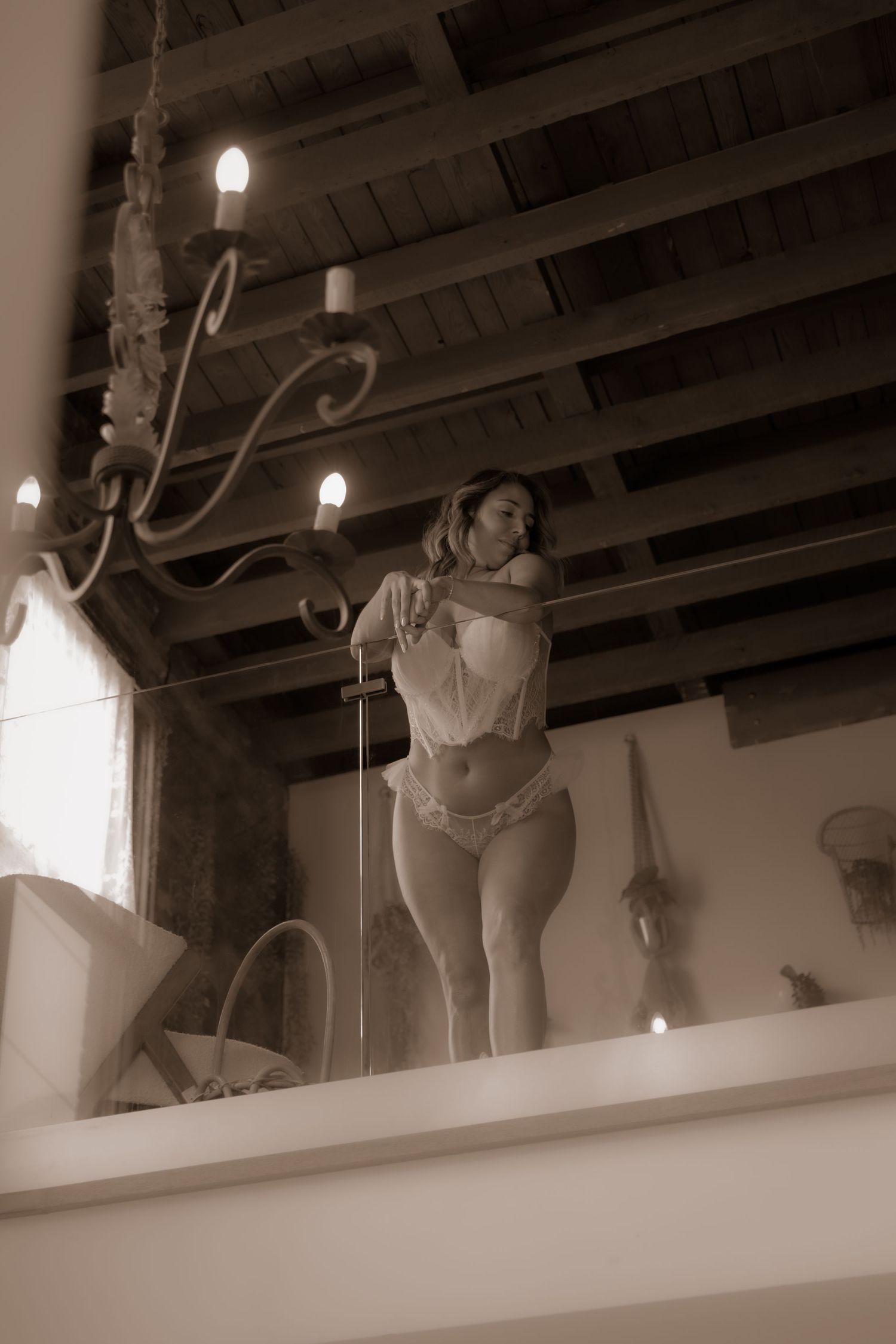 Sepia-toned boudoir photograph under vintage chandelier in rustic setting.