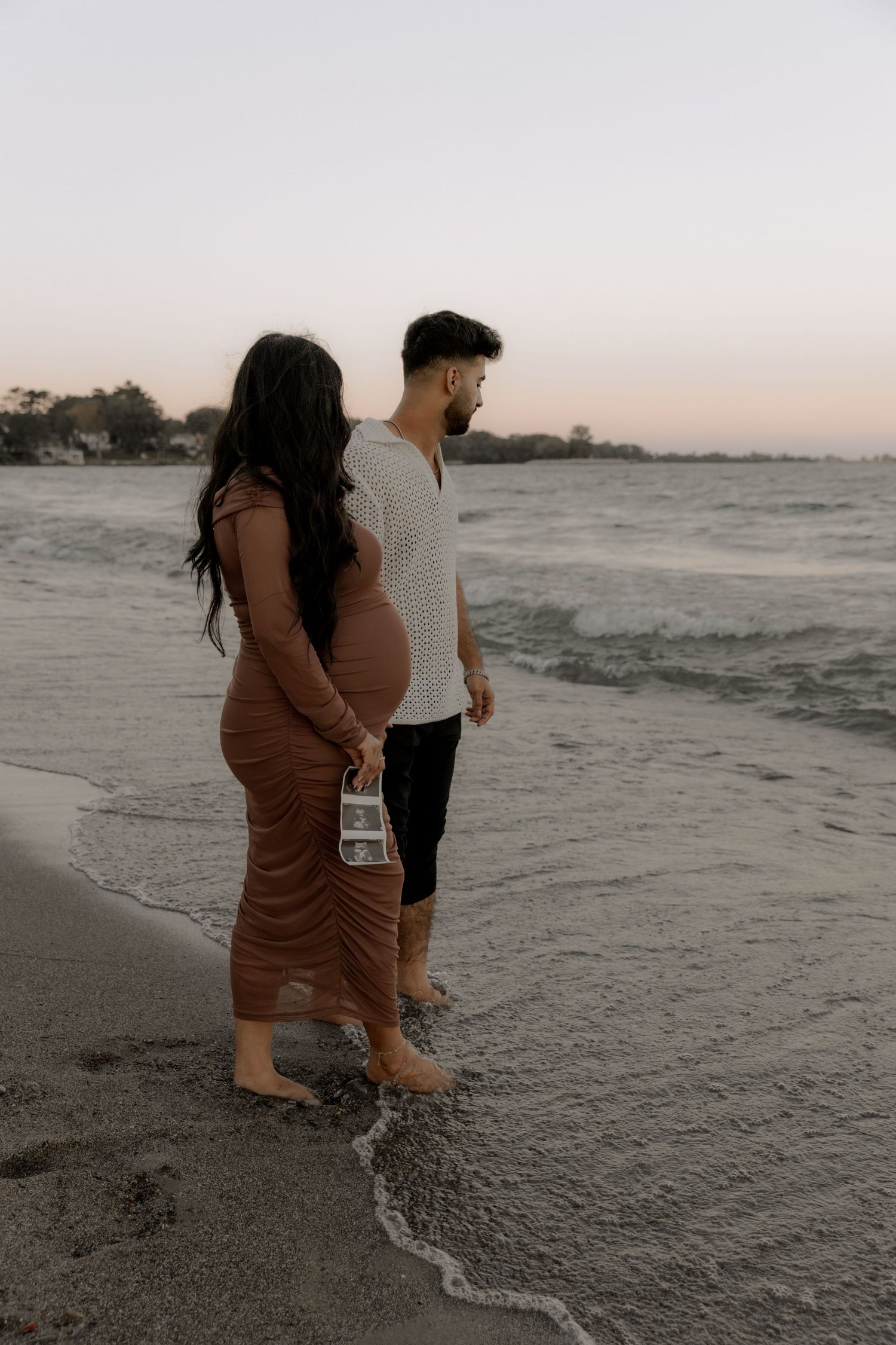 Couple walking along shoreline at sunset with pregnancy ultrasound photos.