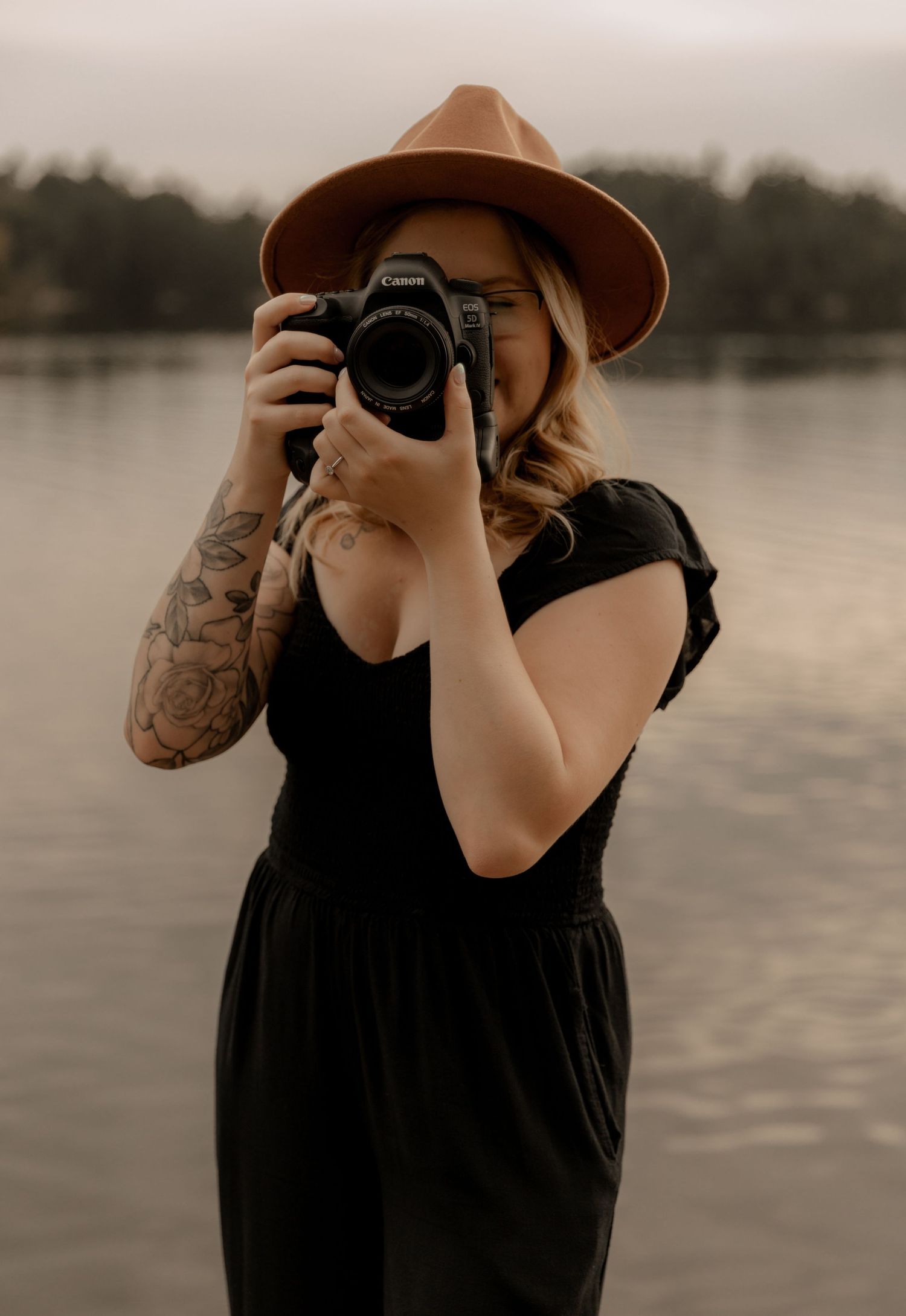 Person with tattoos takes photos with a DSLR camera while wearing a brown hat.