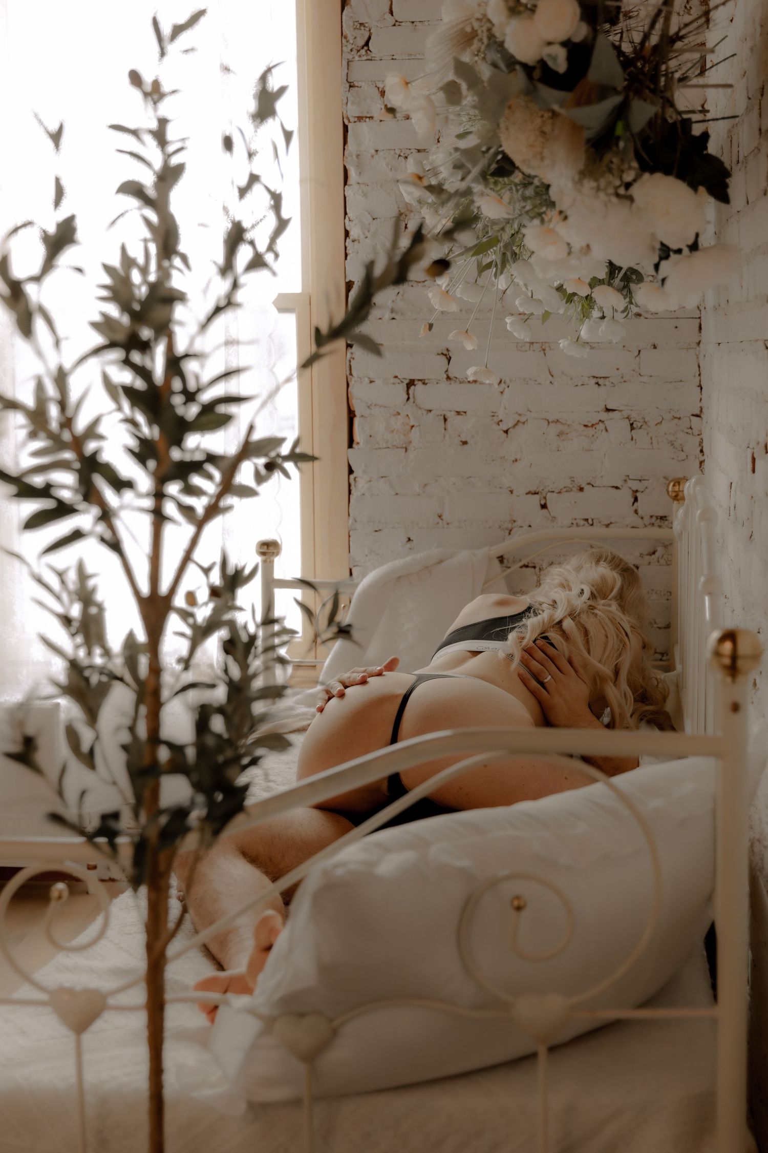 Moody boudoir photography featuring simple white bedding against weathered brick with natural plant elements.