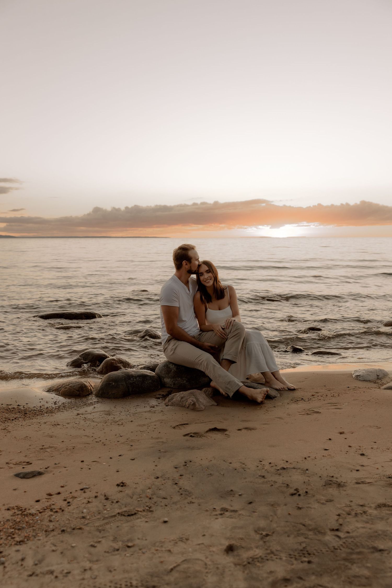 Romantic couple sits on rocky beach during golden sunset over ocean waves.