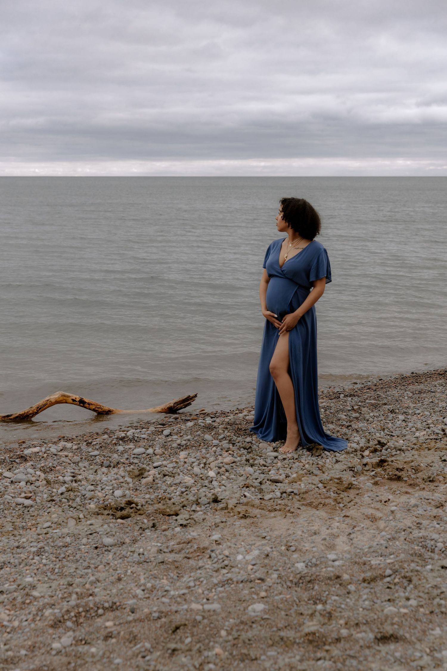 Pregnant woman in blue dress standing on rocky beach holding belly, overcast sky and calm ocean in background.