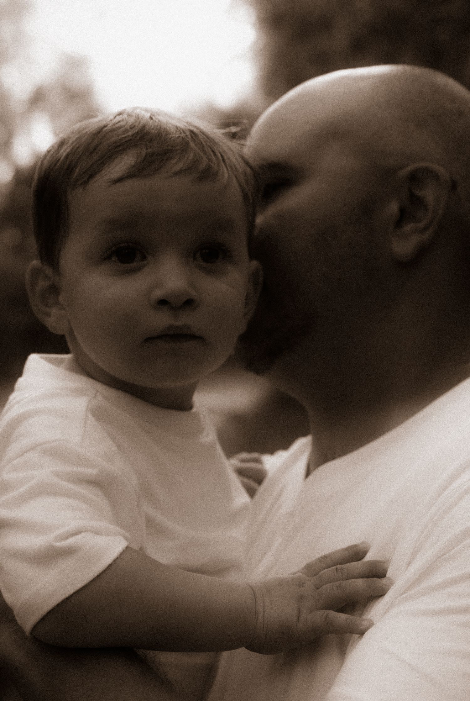 A bald father tenderly kisses his young toddler son on the cheek while holding him in a sepia-toned portrait.