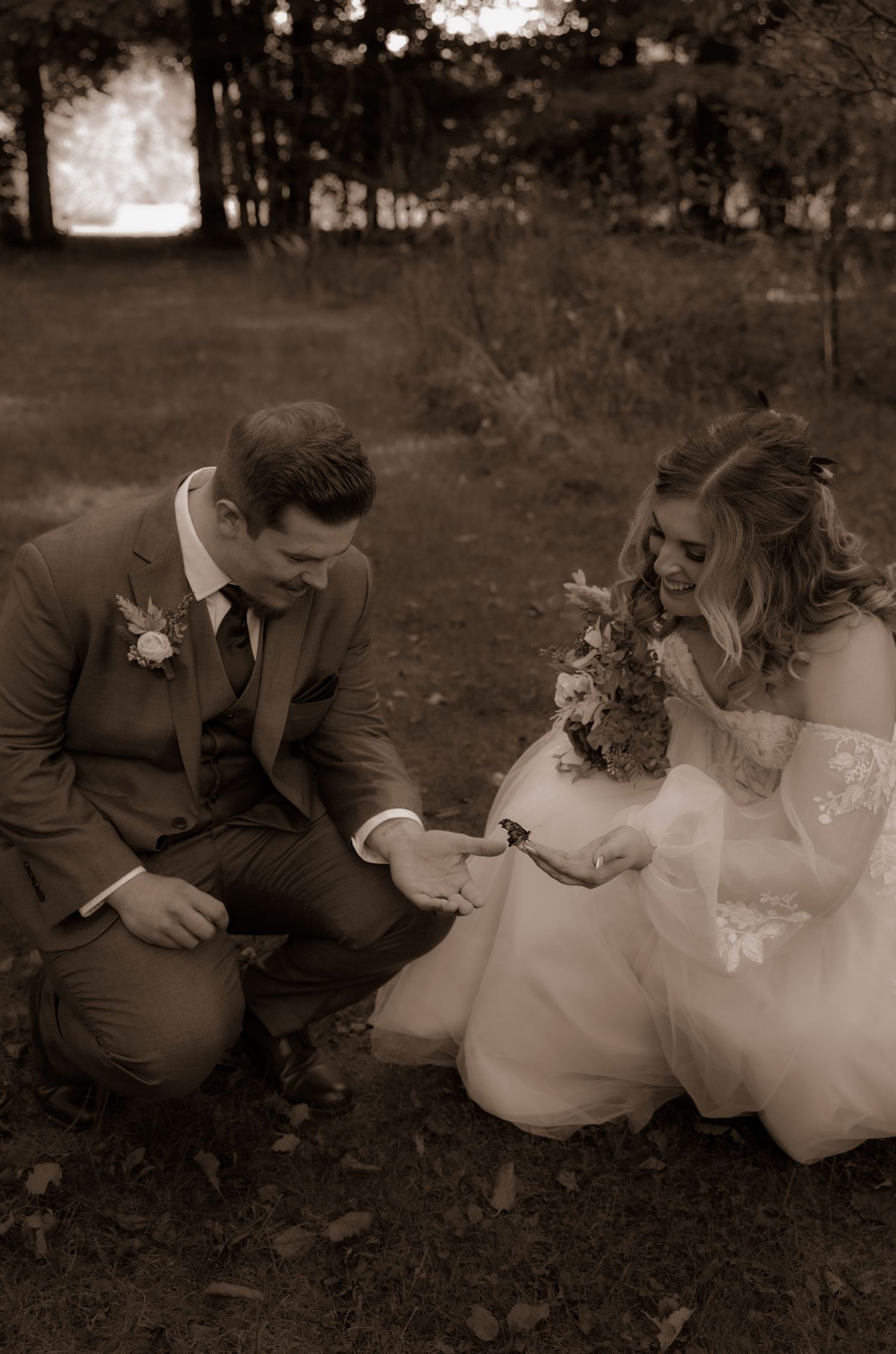 A wedding photo of a couple in formal attire kneeling together outdoors in a wooded setting.