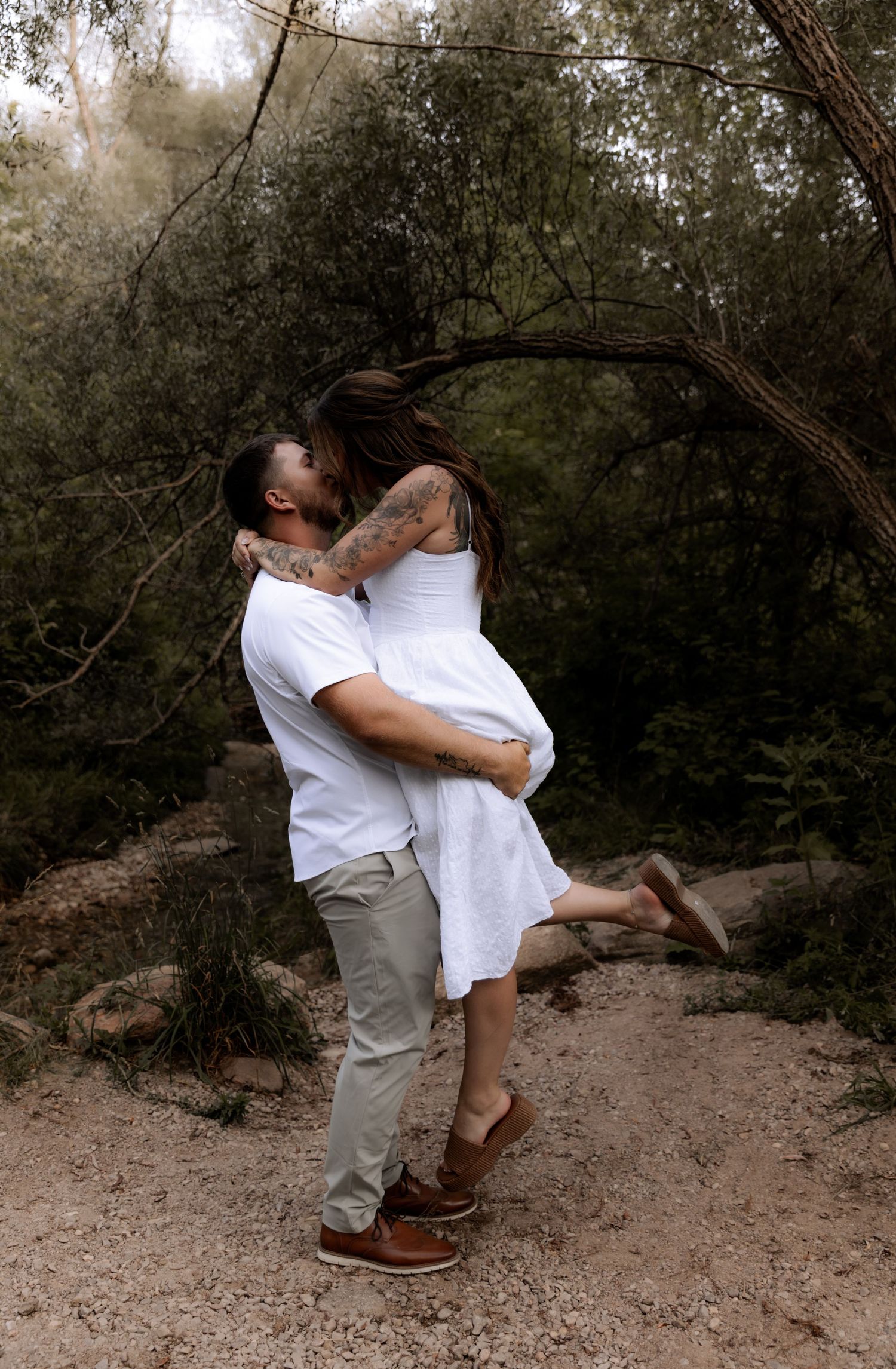 A couple shares a romantic kiss as the man lifts the woman in a lush forest setting, both wearing white outfits.