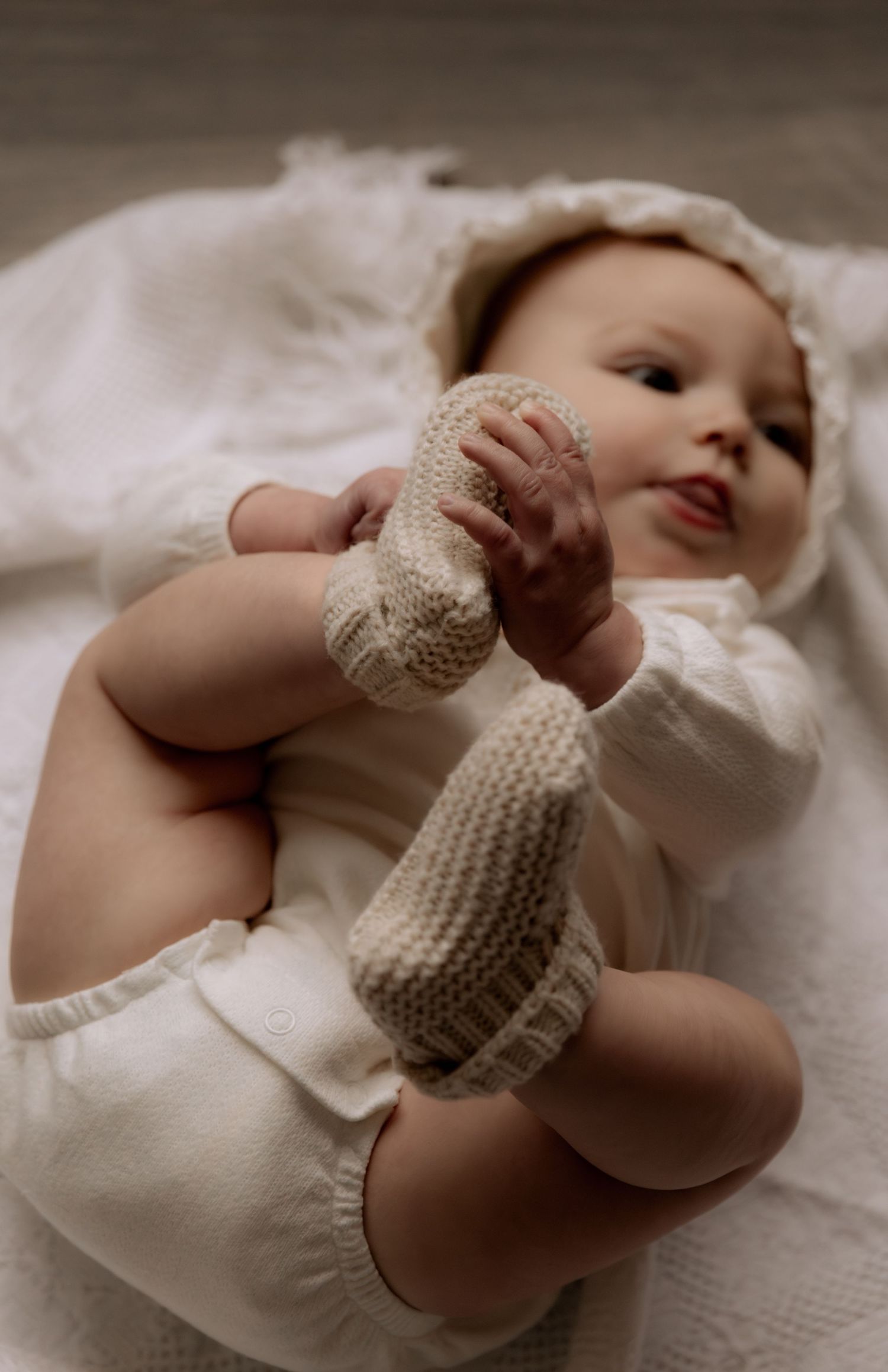 Adorable baby in white outfit and knit booties lying on a soft blanket, playfully holding their feet.