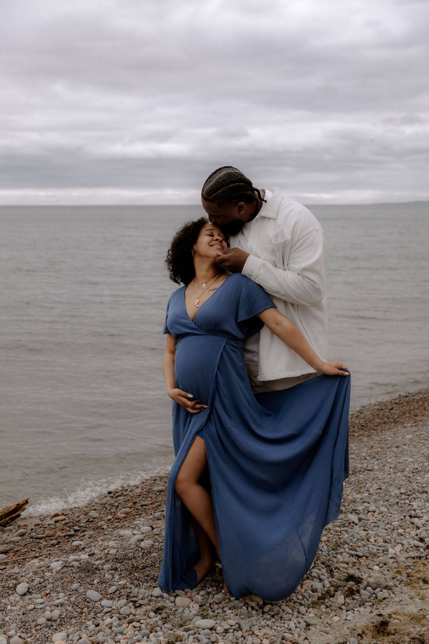 Pregnant woman in blue dress with partner on rocky beach, sharing tender moment with overcast sky and calm ocean backdrop.