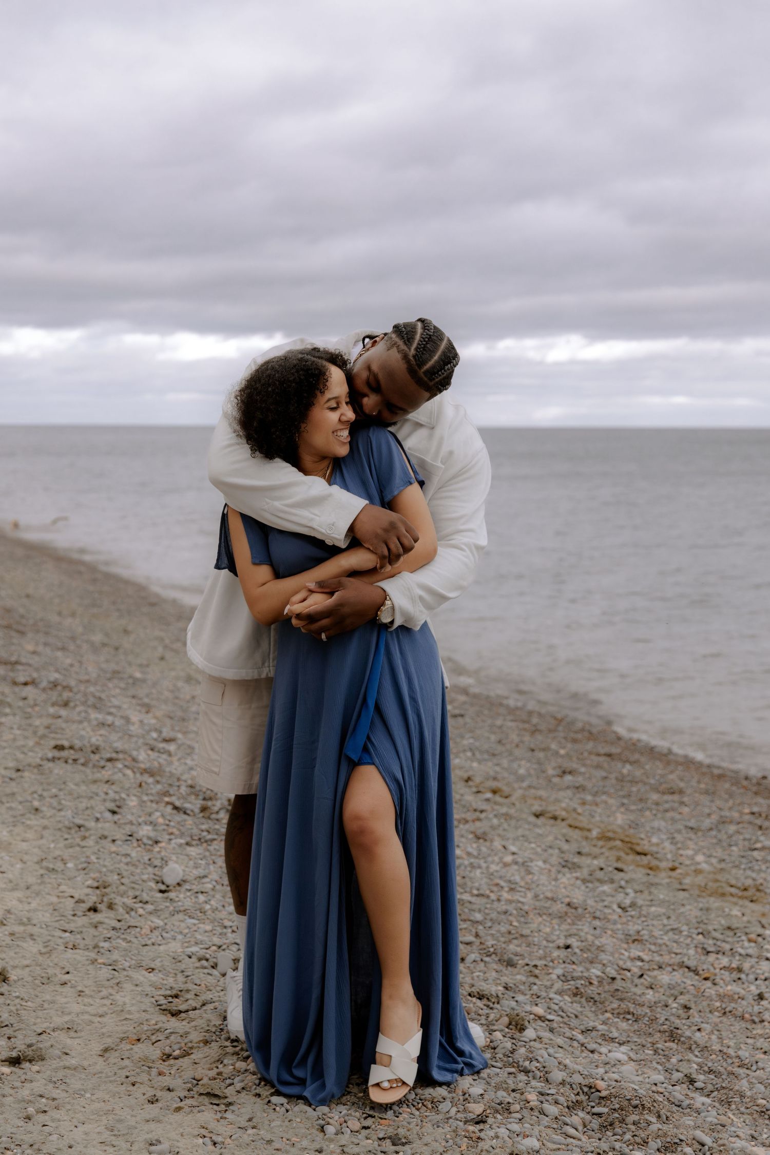 Couple embracing on a rocky beach, woman in blue maxi dress, man in white shirt, overcast sky and ocean backdrop.