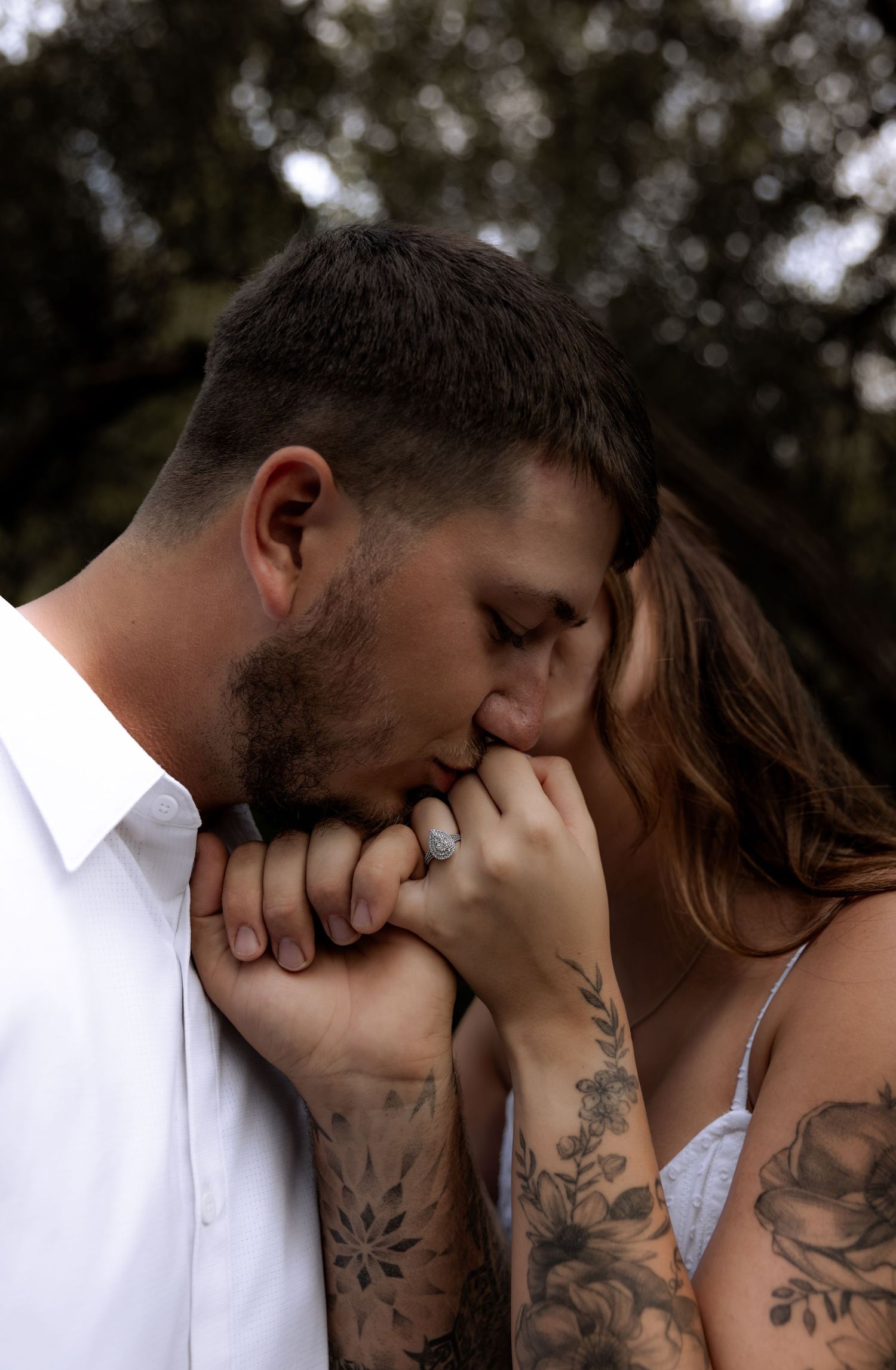 A tattooed couple shares a romantic moment as the man kisses the woman's hand, showcasing her diamond engagement ring.