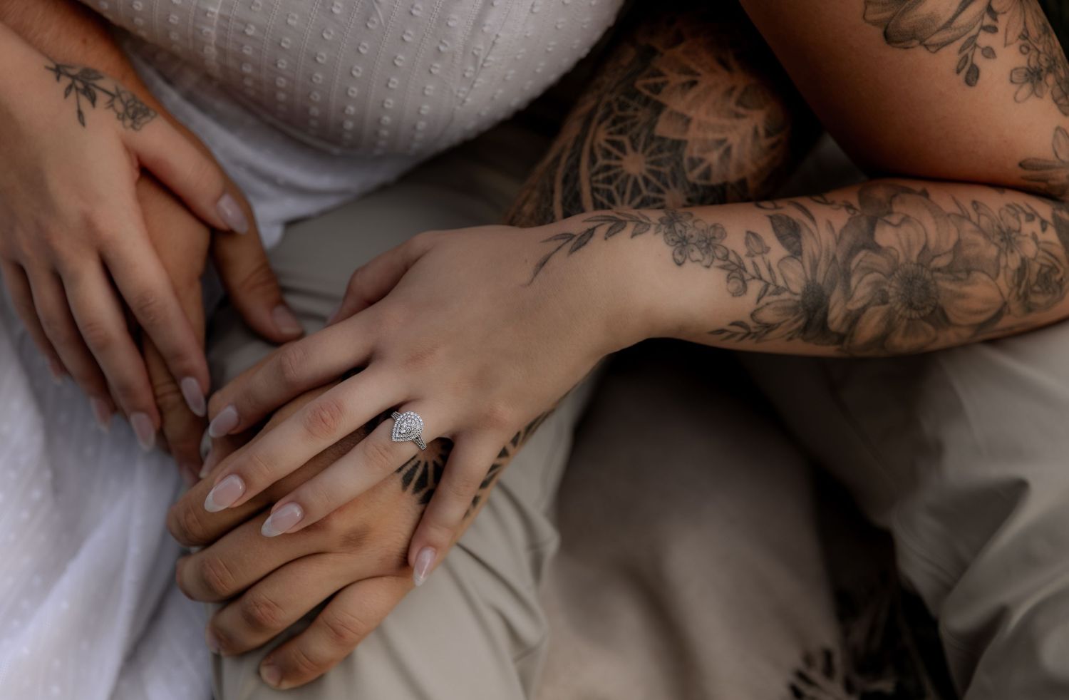 Close-up of intertwined hands with tattoos and a diamond engagement ring resting on a white blanket.