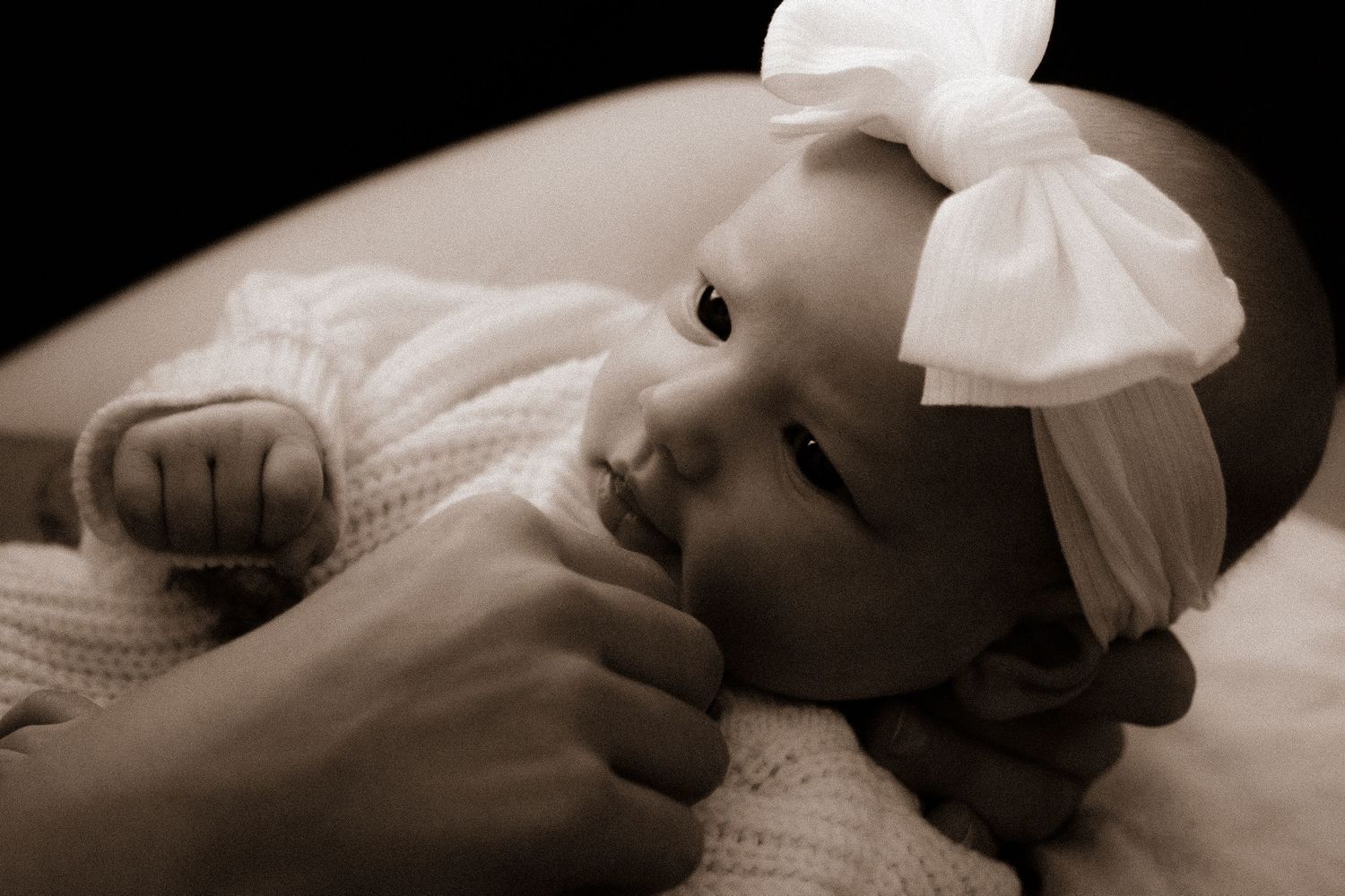 Sepia-toned newborn baby girl in white knit outfit and bow headband, gently held by an adult's hands.