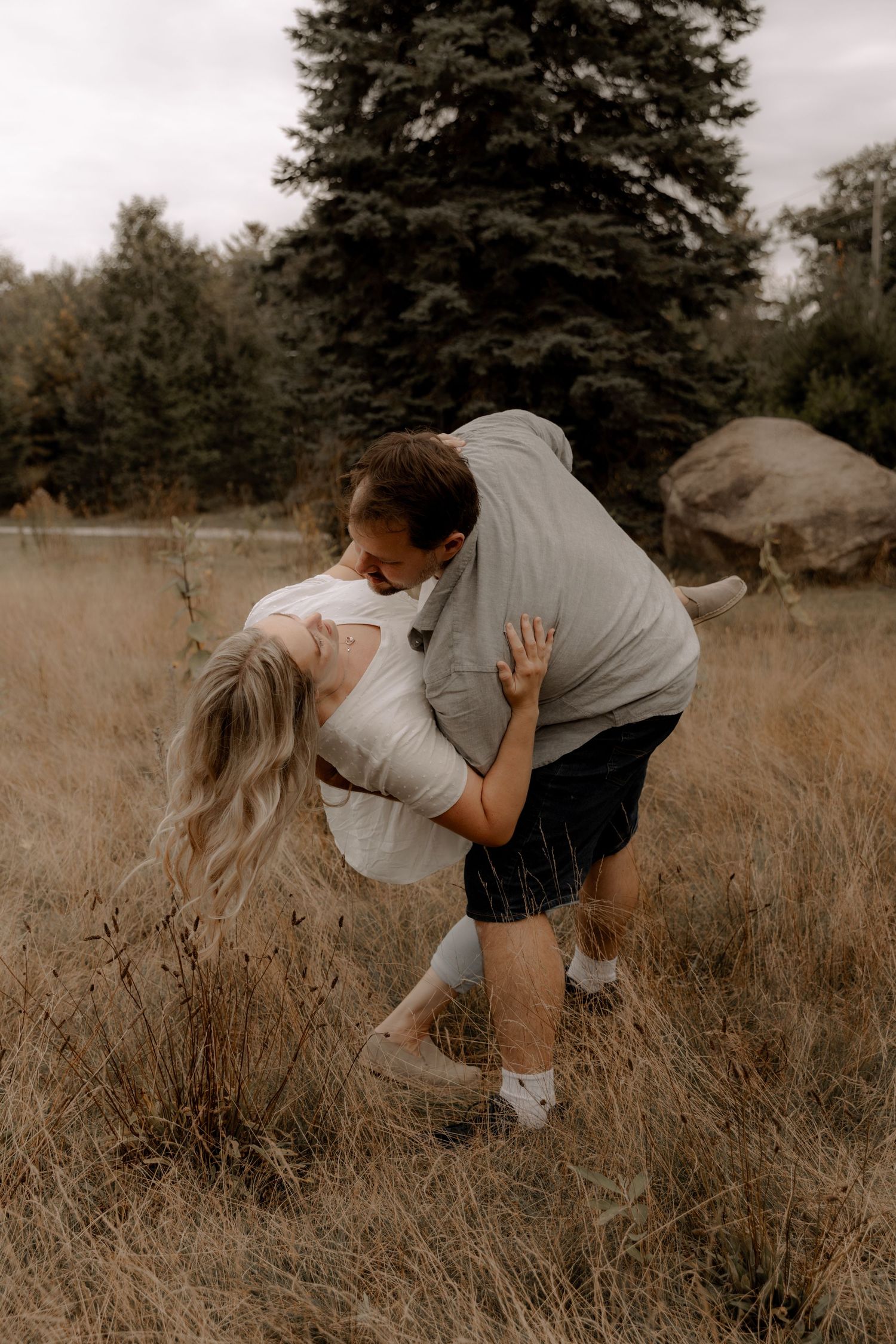 Dancing couple shares romantic dip in rustic field with pine trees.