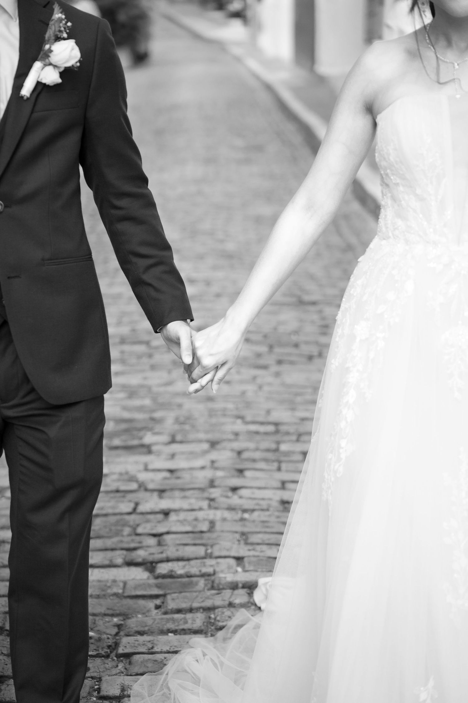 Black and white photo of bride and groom holding hands on cobblestone street, wedding attire visible.
