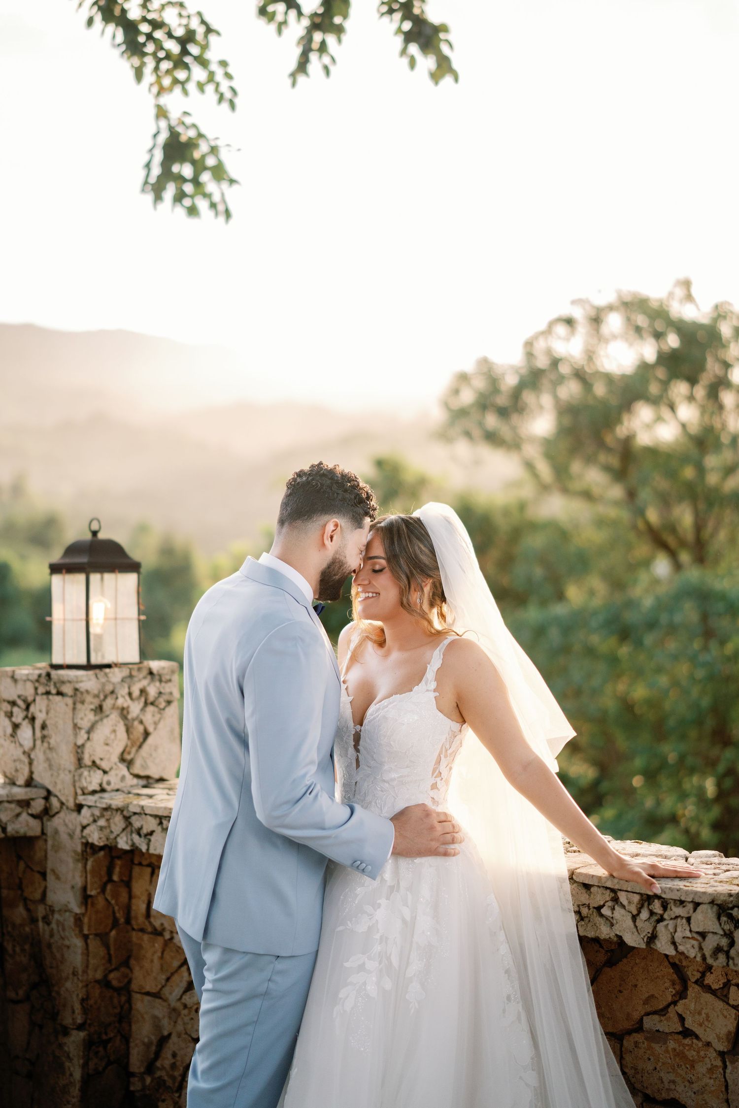Bride and groom share romantic moment at golden hour, embracing on stone terrace with mountain views.