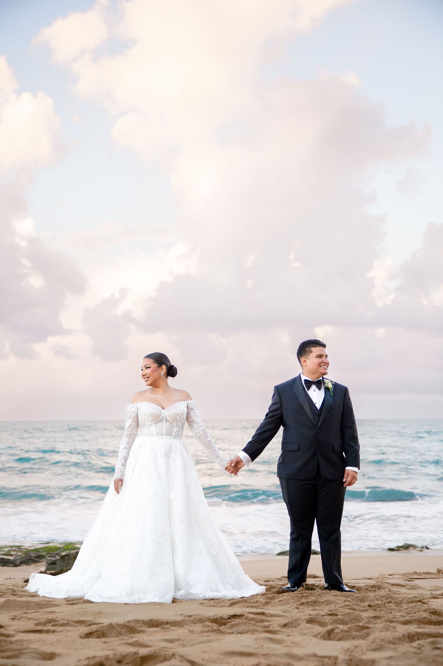Bride and groom holding hands on a sandy beach at sunset, bride in lace gown, groom in black tuxedo.