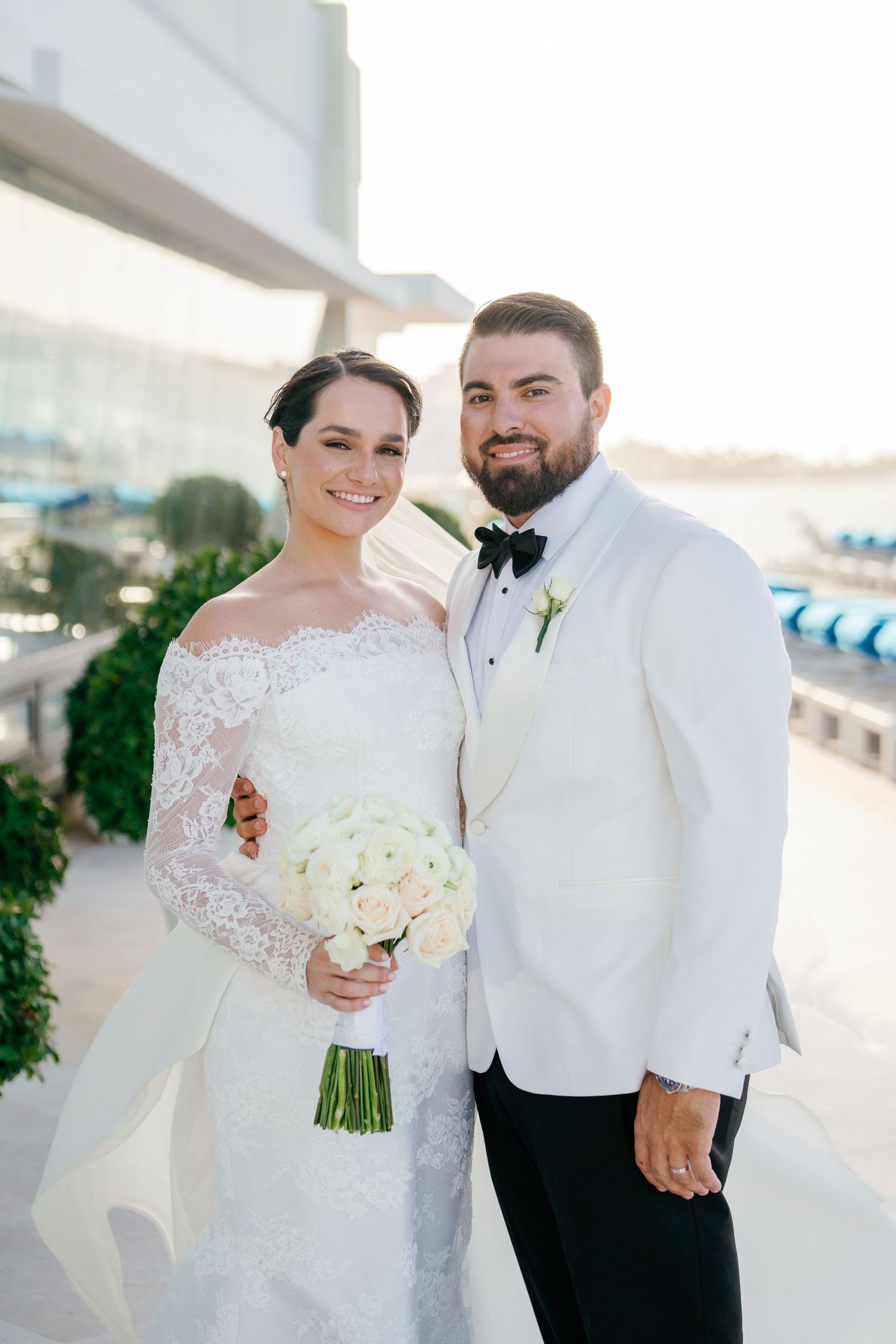 Bride in lace off-shoulder gown holding white bouquet stands with groom in white tuxedo jacket and black bow tie.