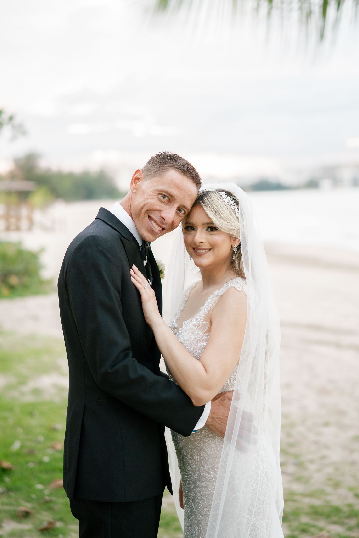 Bride and groom embracing on a tropical beach, smiling at camera, bride in lace gown with veil, groom in black tuxedo.