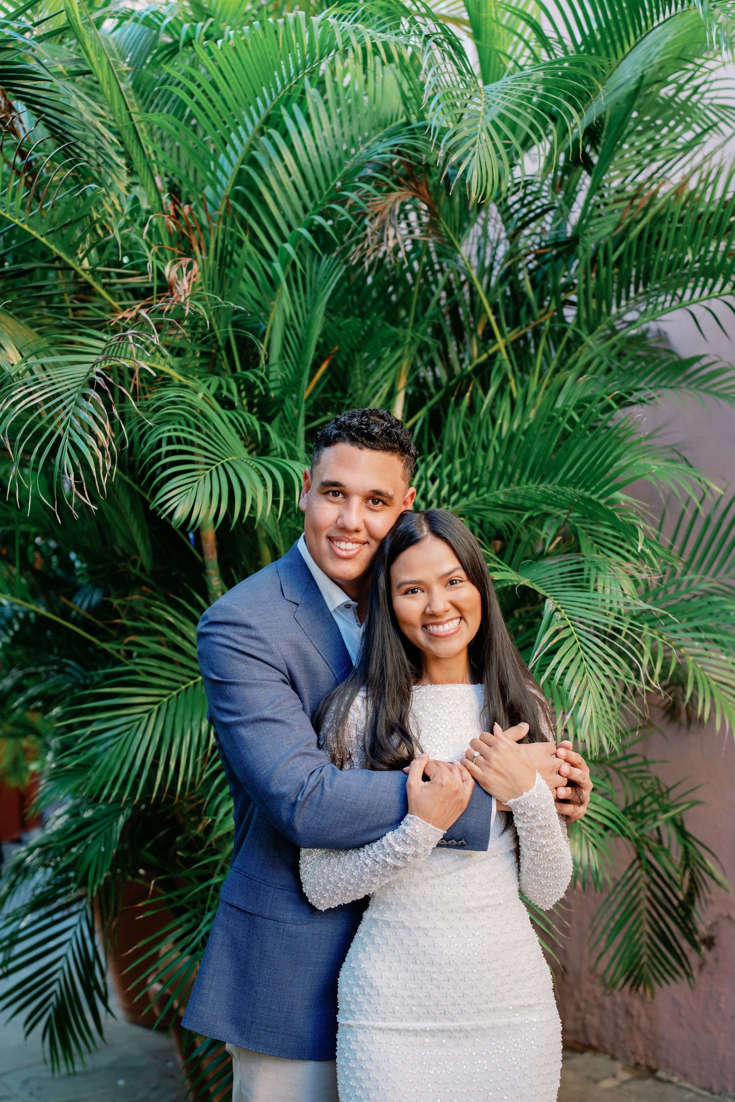 Smiling couple embraces in front of lush tropical palm tree, man in blue suit hugging woman in white dress.