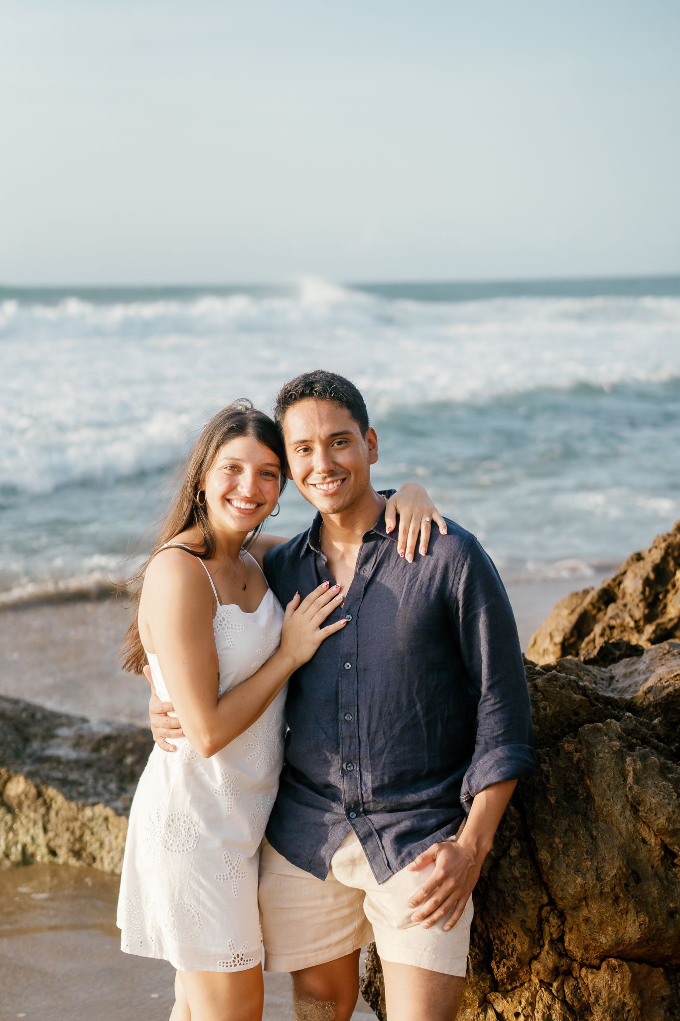A romantic couple embraces on a rocky beach with ocean waves and blue sky in the background.