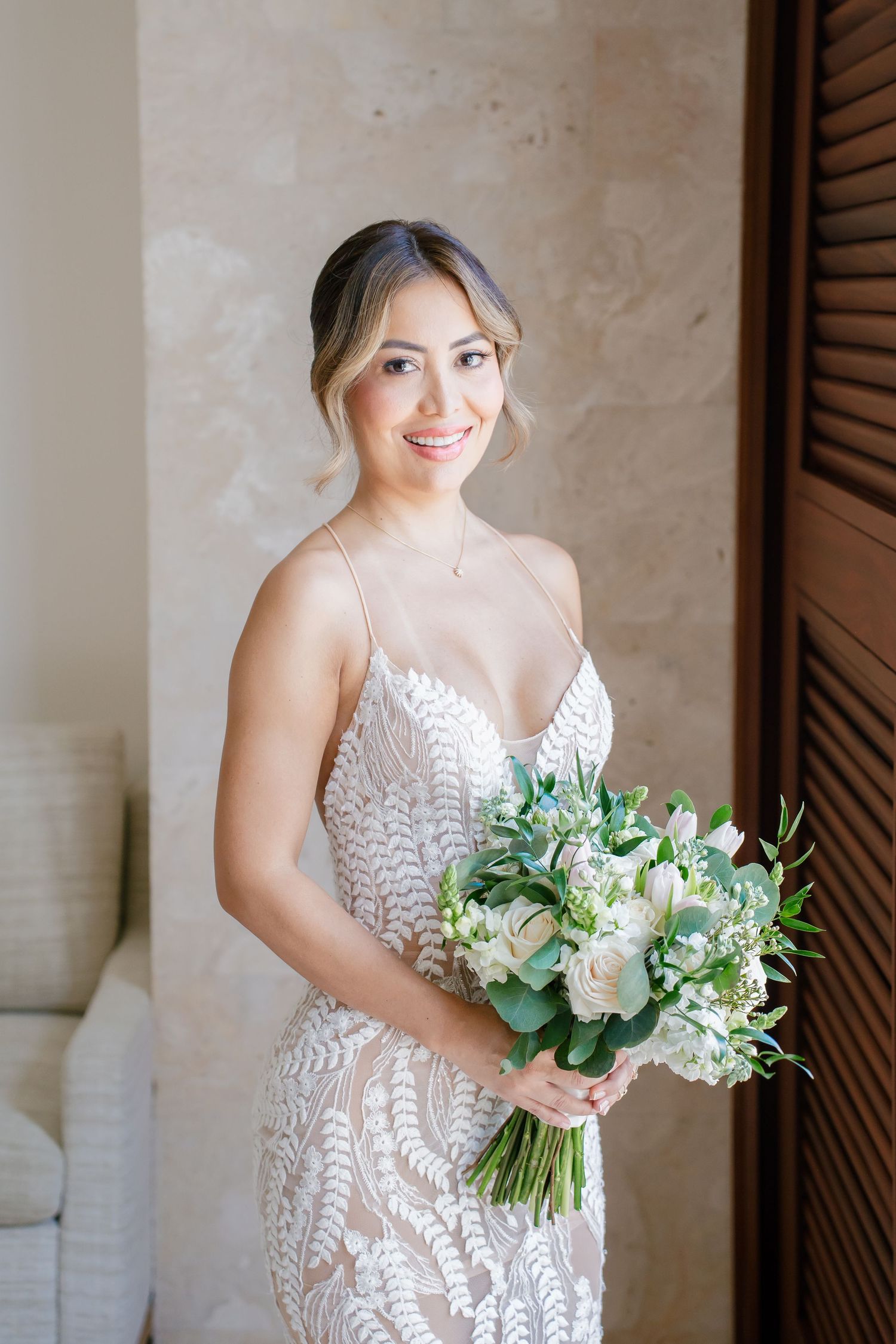 Bride wearing lace wedding gown holds beautiful white and green floral bouquet near wooden shutters.