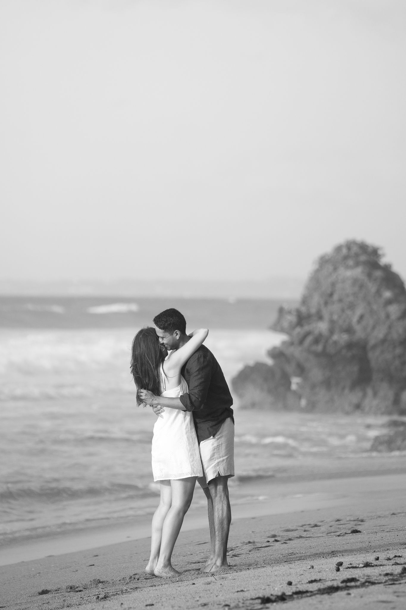 A couple shares a romantic moment walking together along a misty beach shoreline in black and white.