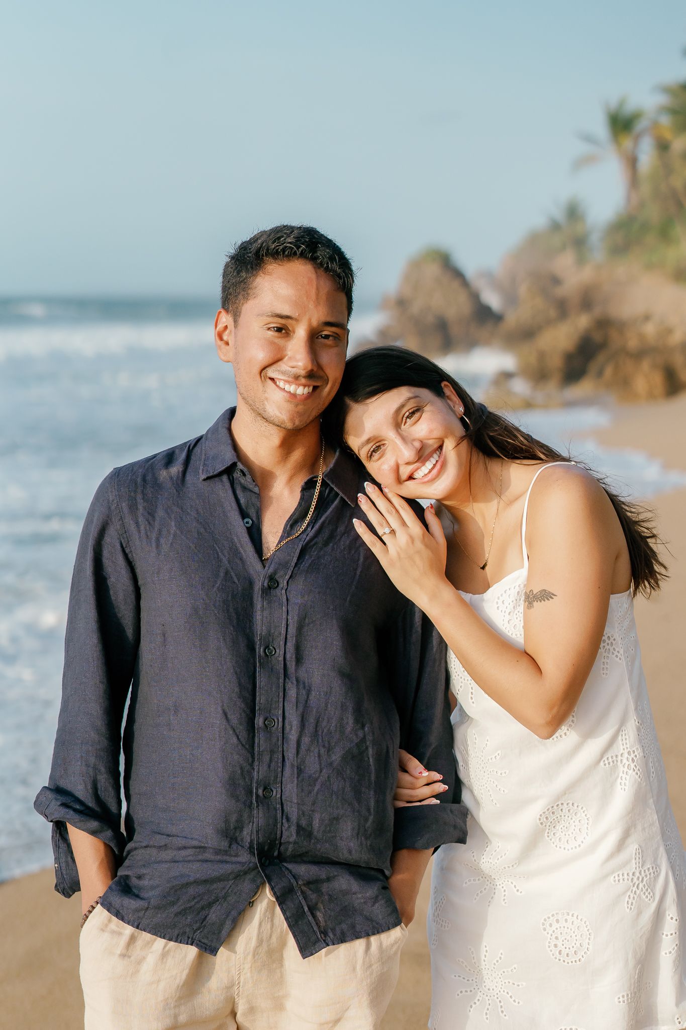 A couple shares joyful moments together on a sunny beach with palm trees and ocean waves in the background.