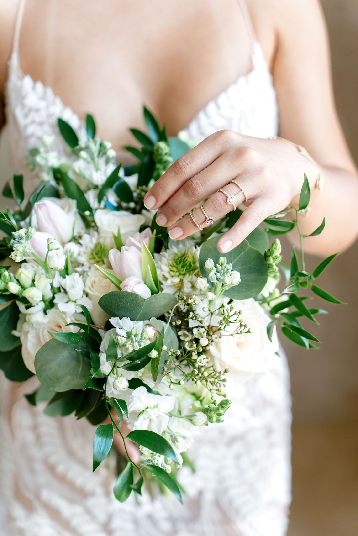 Close up of a bridal bouquet with white flowers and green foliage, showing wedding rings on bride's hand.