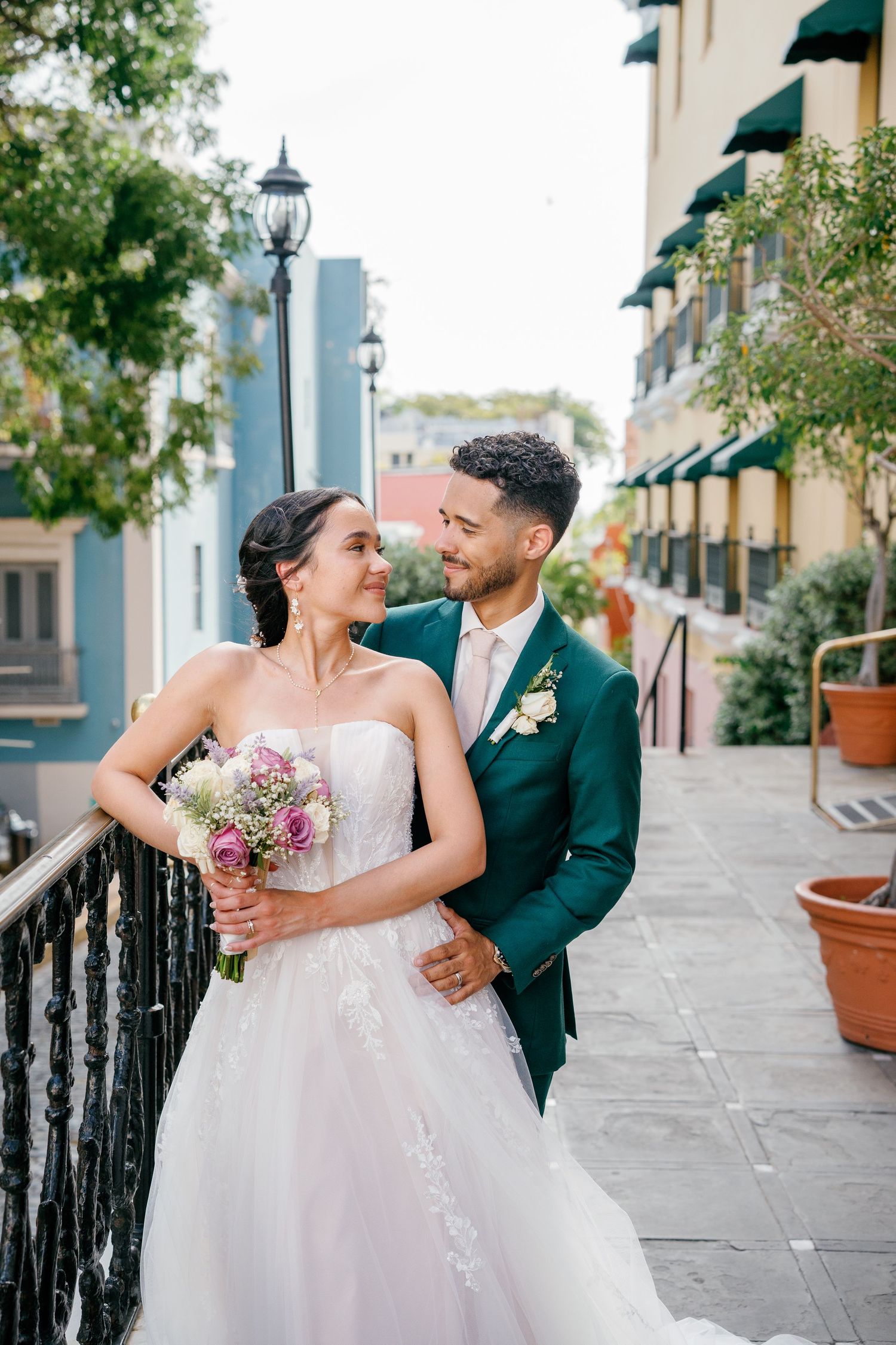 Bride and groom pose on a balcony, she holds a pink bouquet in a white gown, he wears a green suit.