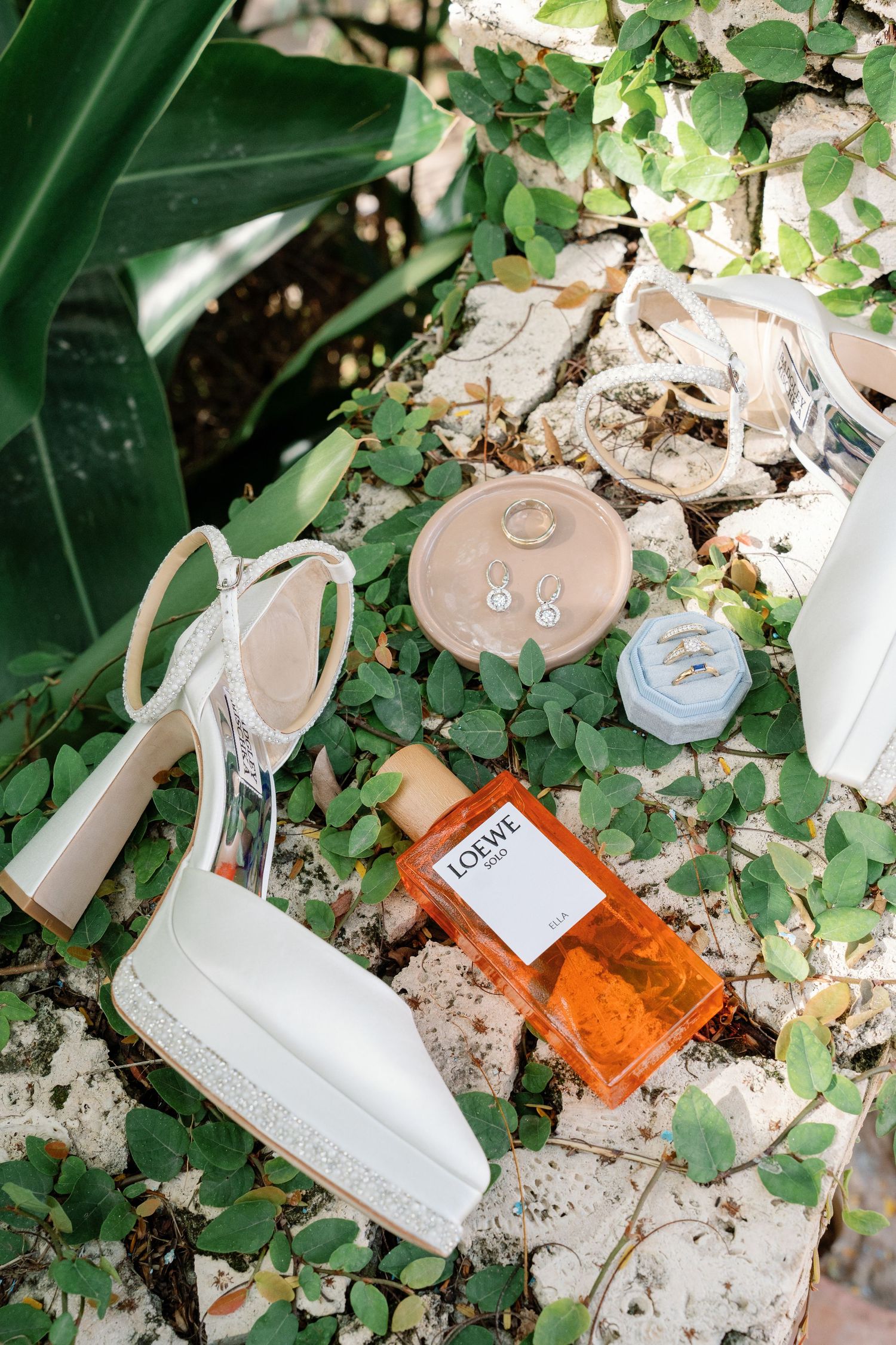 White bridal heels, Loewe perfume, earrings, and ring boxes arranged on stone surface with green ivy leaves.