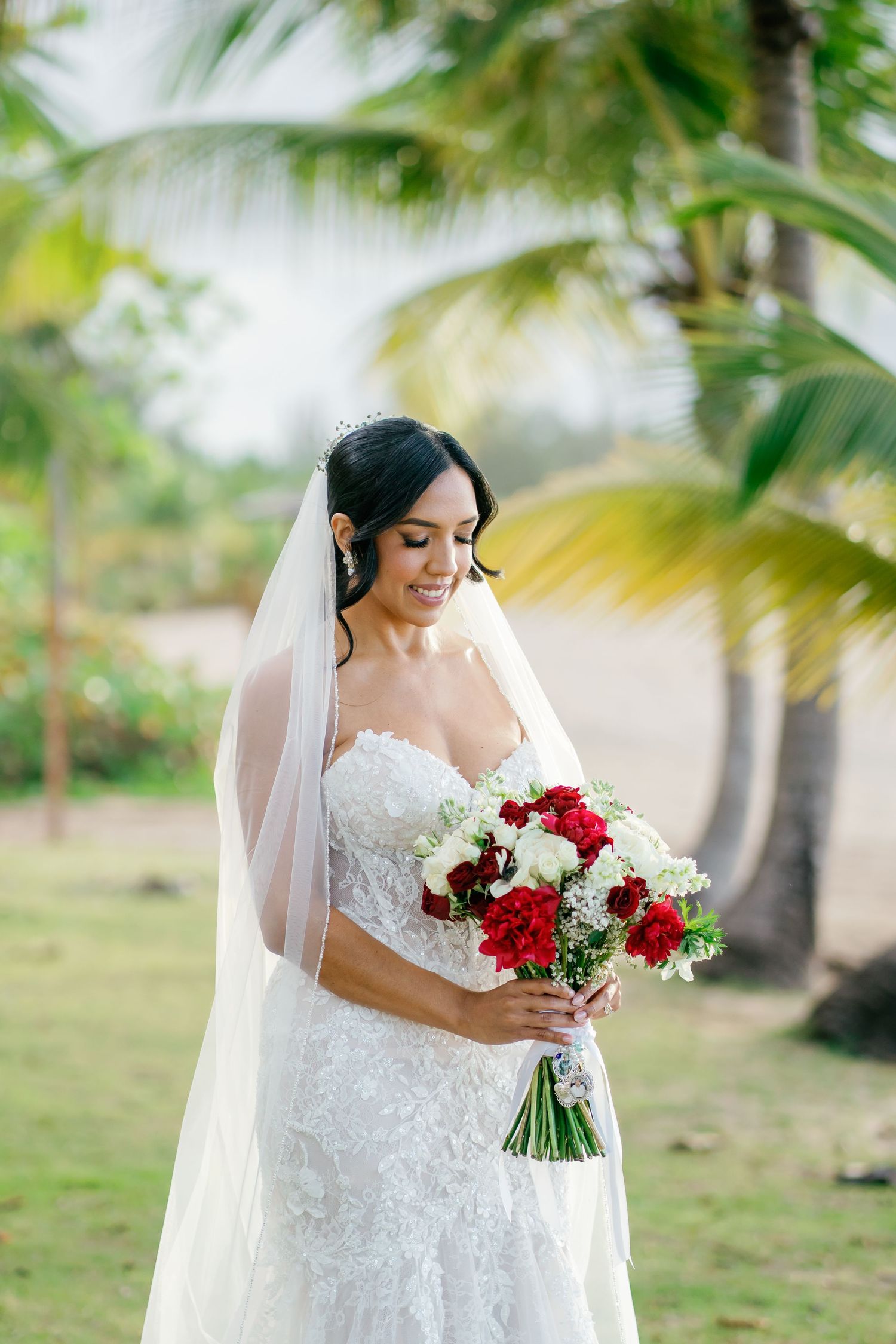 Smiling bride in lace wedding gown holding red and white bouquet beneath tropical palm trees.