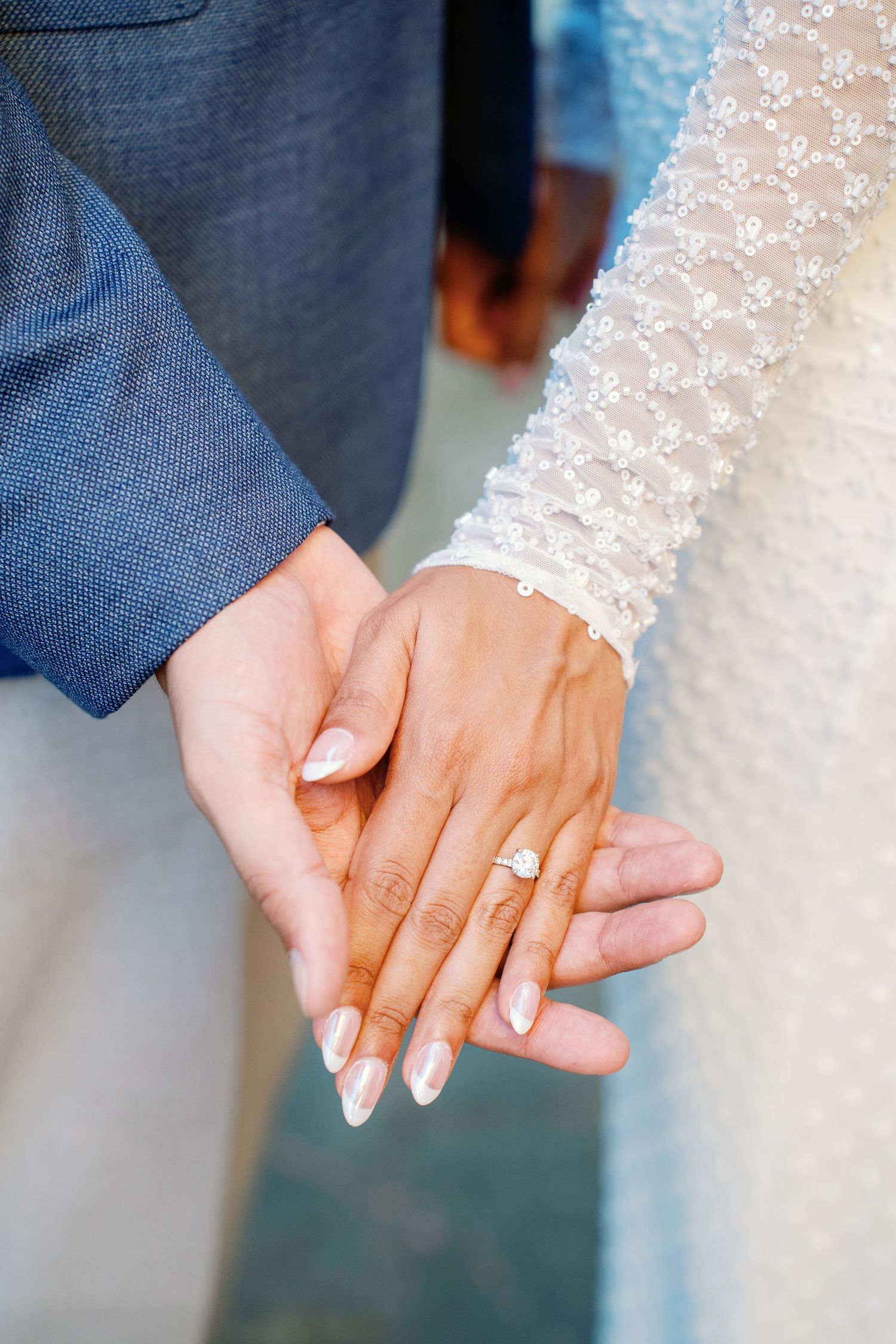 Newlywed couple holding hands, bride wearing diamond engagement ring and lace wedding dress sleeve, groom in blue suit.