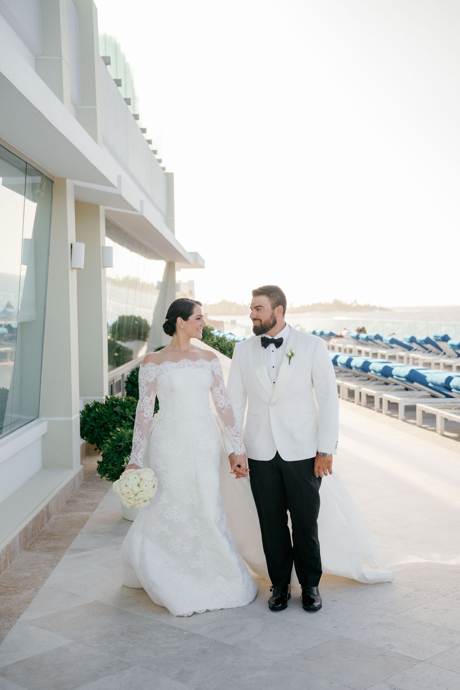 Bride and groom hold hands walking on a seaside terrace, she in a lace gown, he in a white tuxedo jacket.
