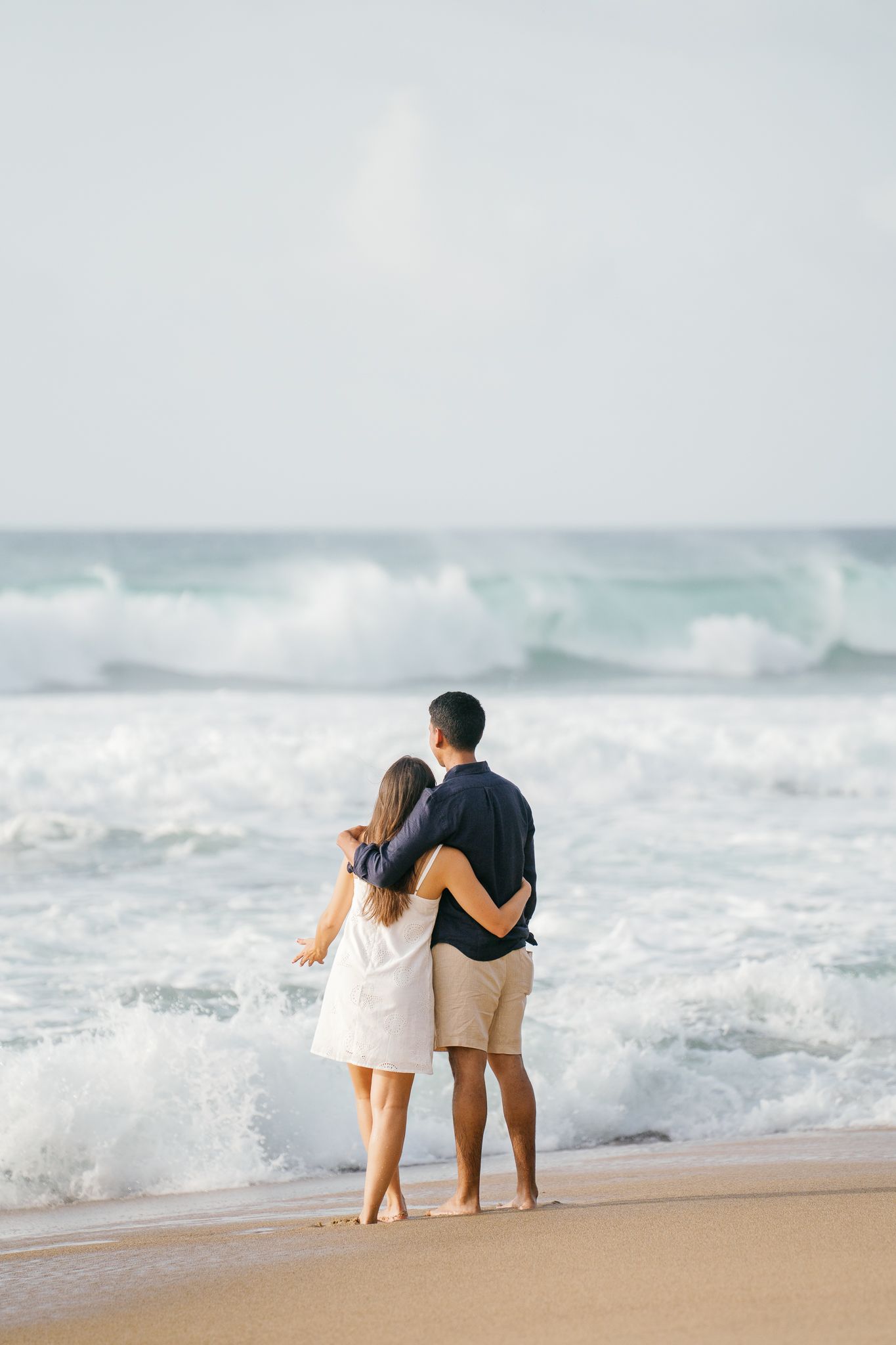 A couple stands together on a sandy beach while waves crash behind them in a romantic coastal scene.