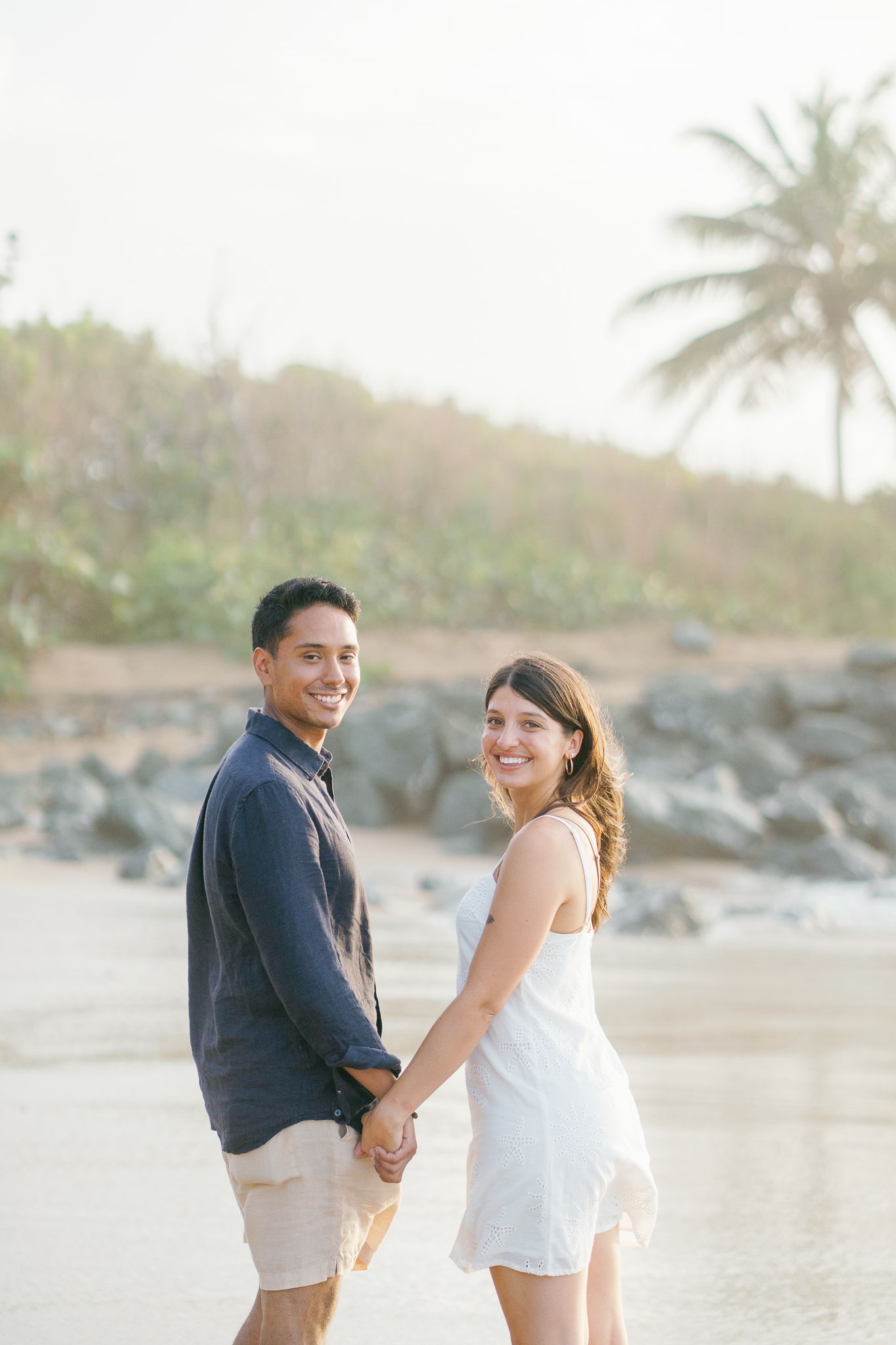 A couple walks along a tropical beach with palm trees and rocks in the background during sunset.