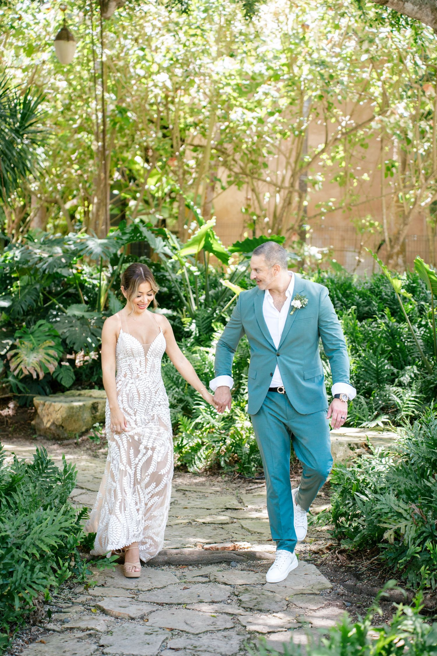 A couple walks hand in hand along a stone garden path surrounded by lush greenery and flowering plants.