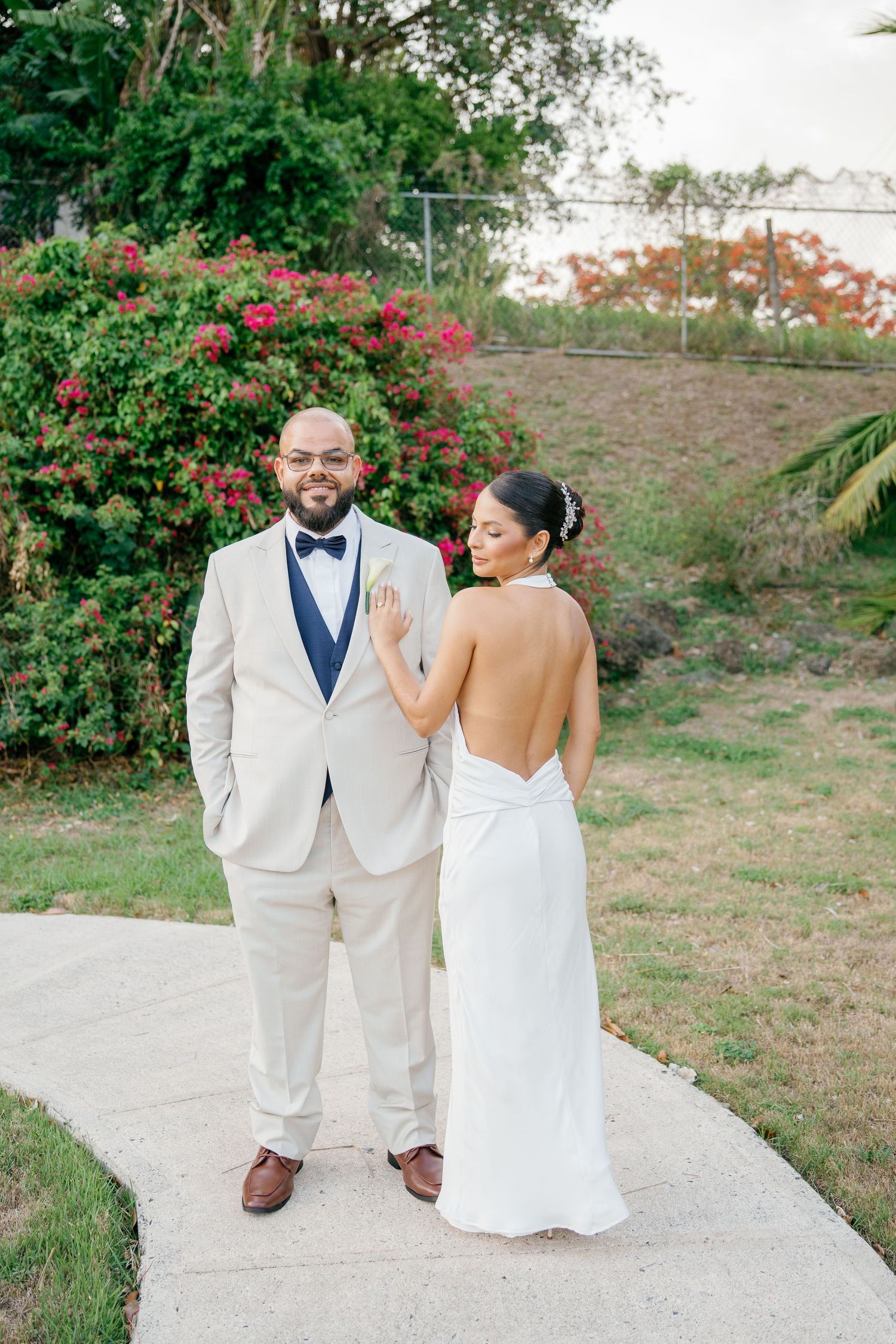 Bride in backless white gown and groom in ivory tuxedo with navy bow tie pose on garden path with tropical flowers.