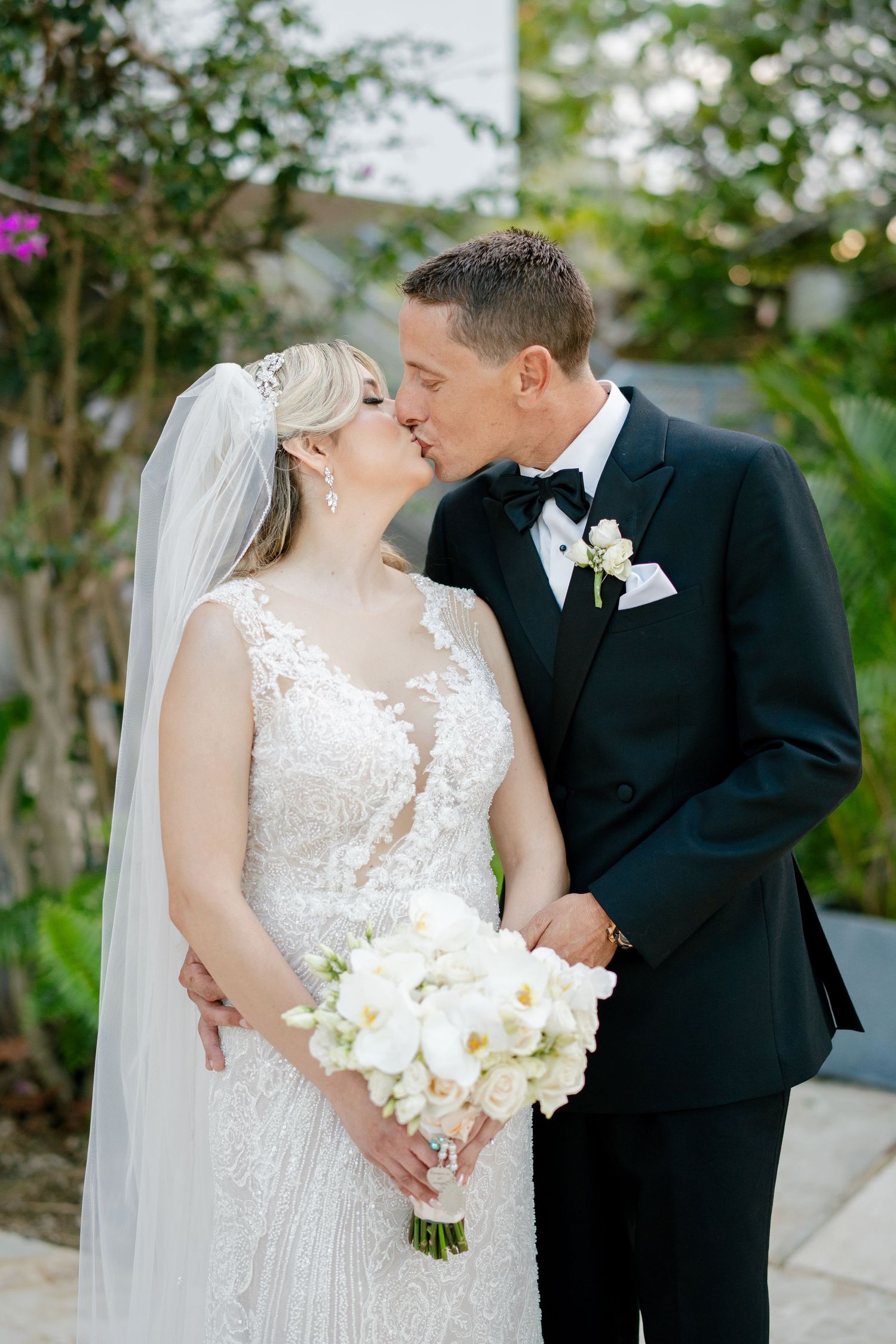 Bride and groom share a romantic kiss, she holds white orchid bouquet, wearing lace gown with veil, he wears black tuxedo.
