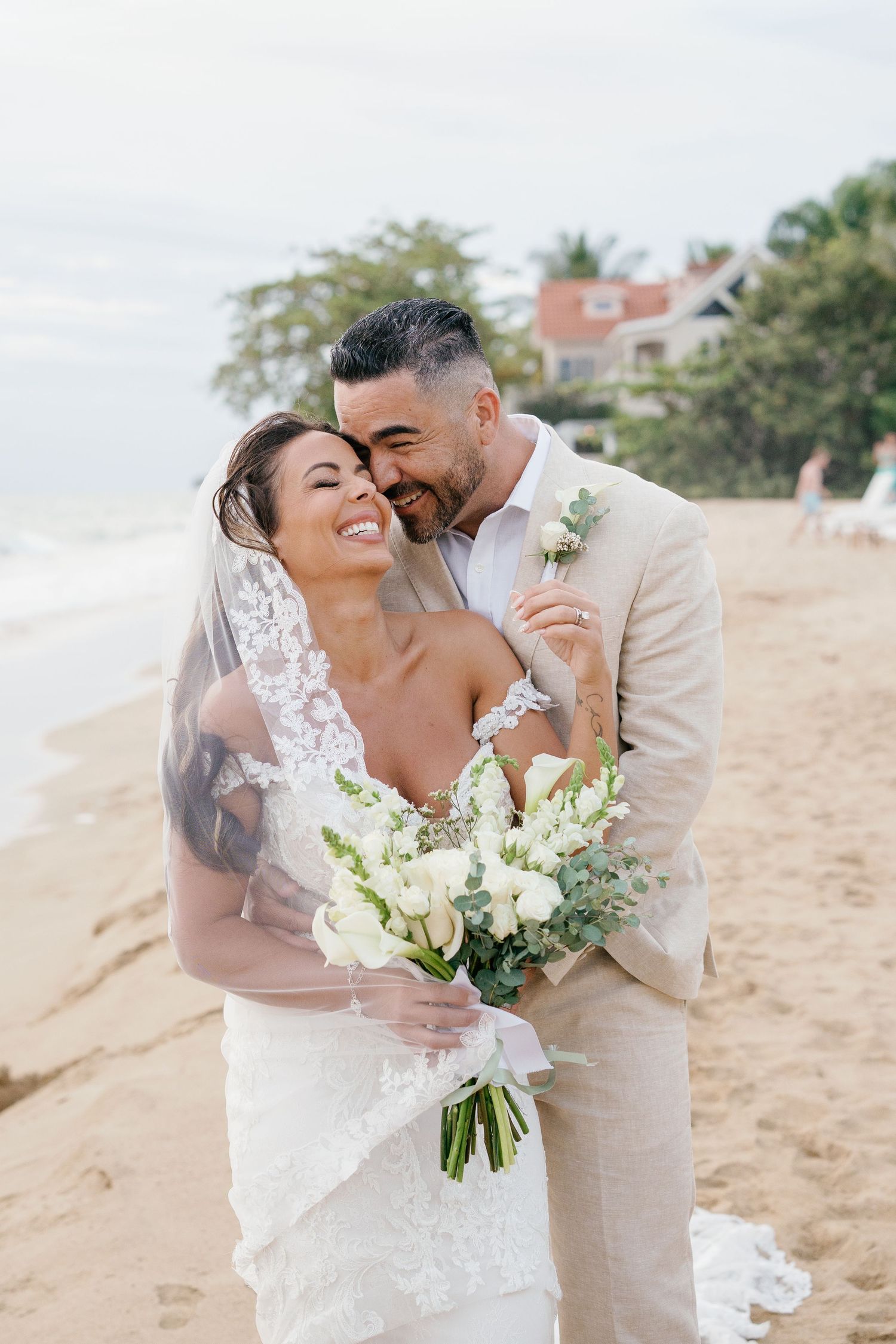 Bride and groom share a romantic beach moment, she holds white bouquet in lace gown, he wears tan suit.