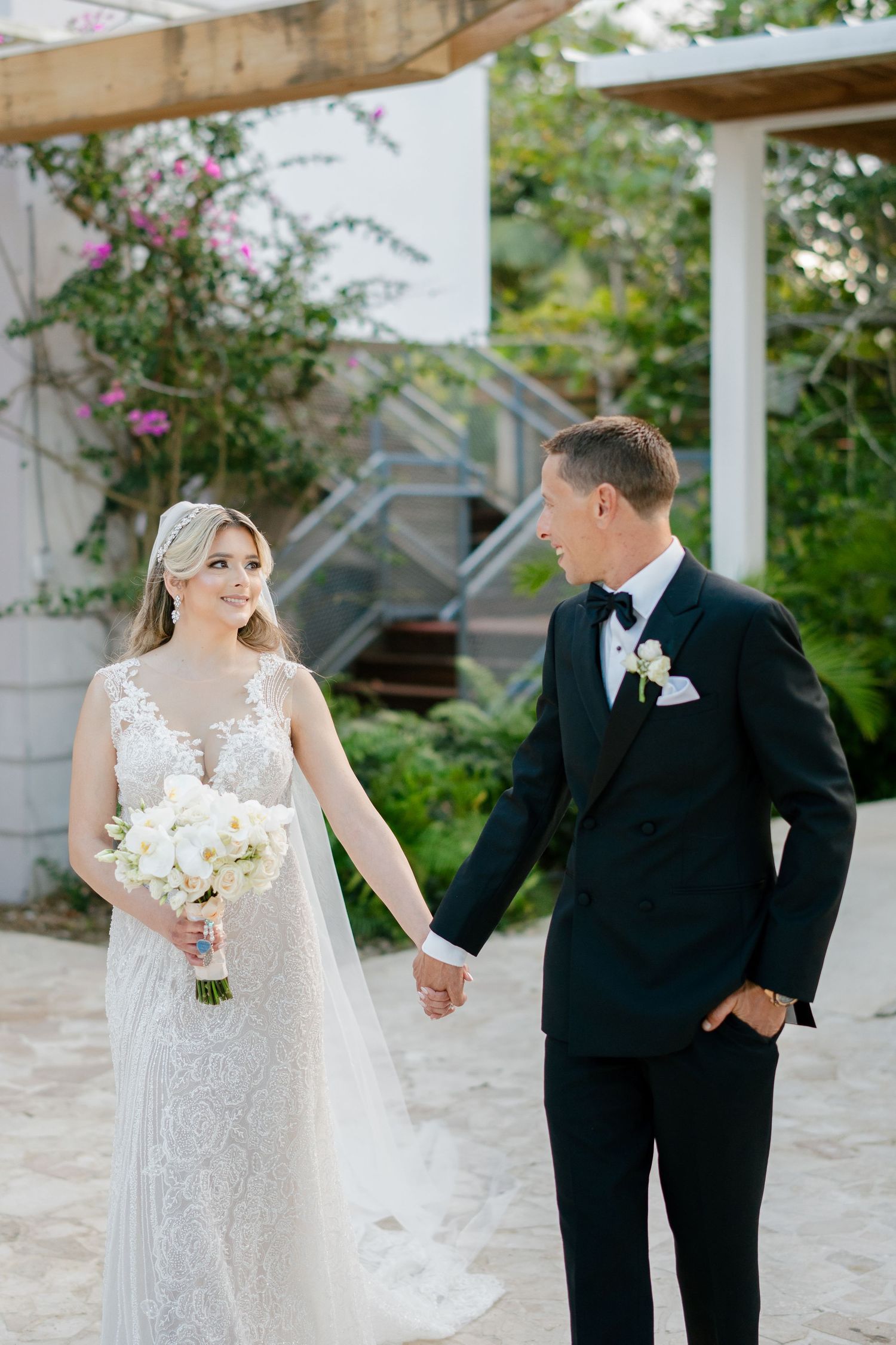 Bride in lace gown holding white bouquet and groom in black tuxedo hold hands at outdoor wedding venue.