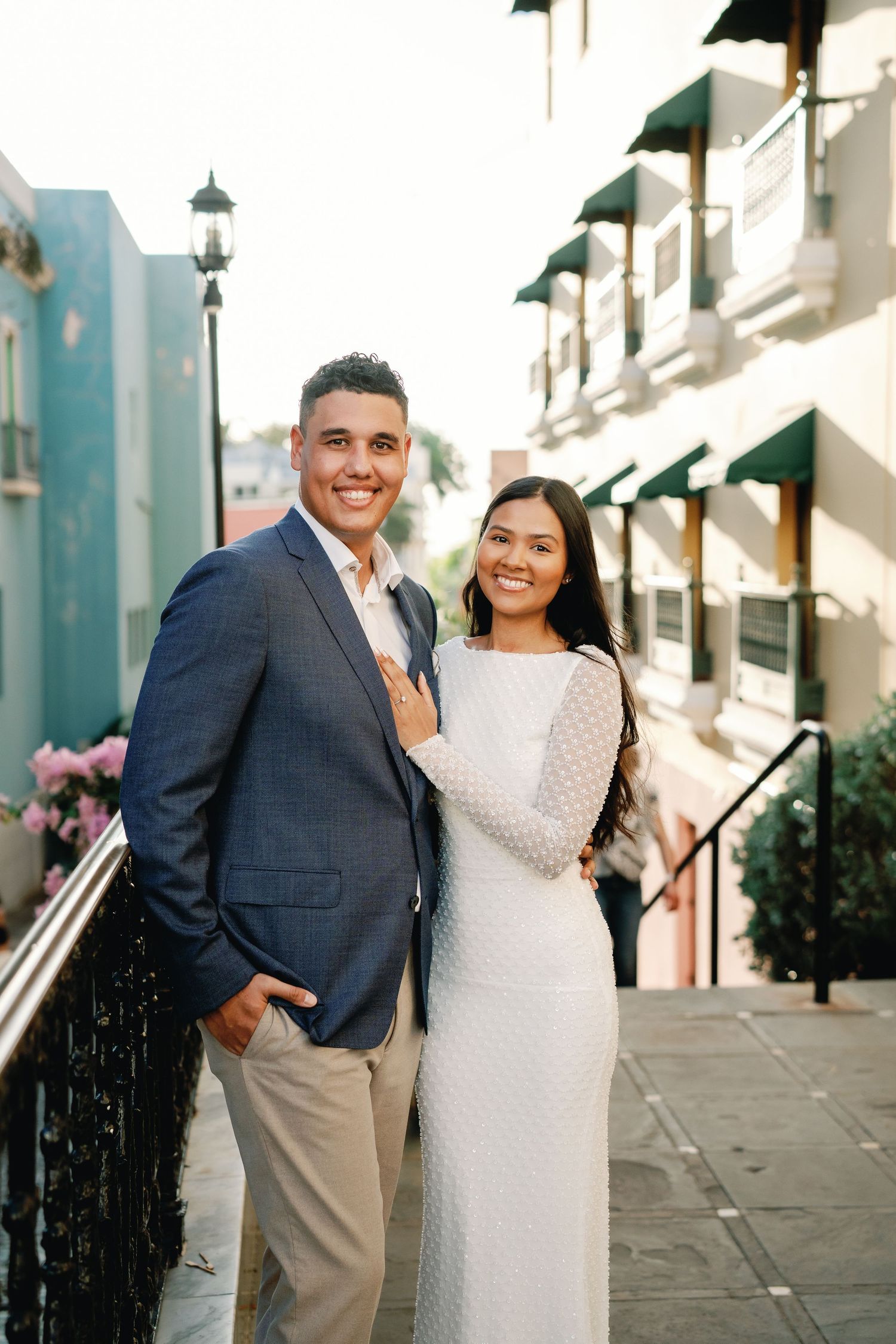 A smiling couple in elegant attire poses on a charming street, the man in a blue blazer and woman in a white lace gown.