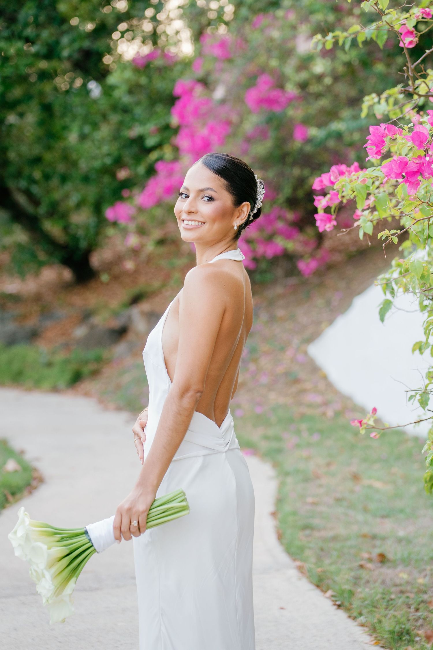Smiling bride in backless white gown holds green calla lily bouquet on garden path with pink bougainvillea blooms.