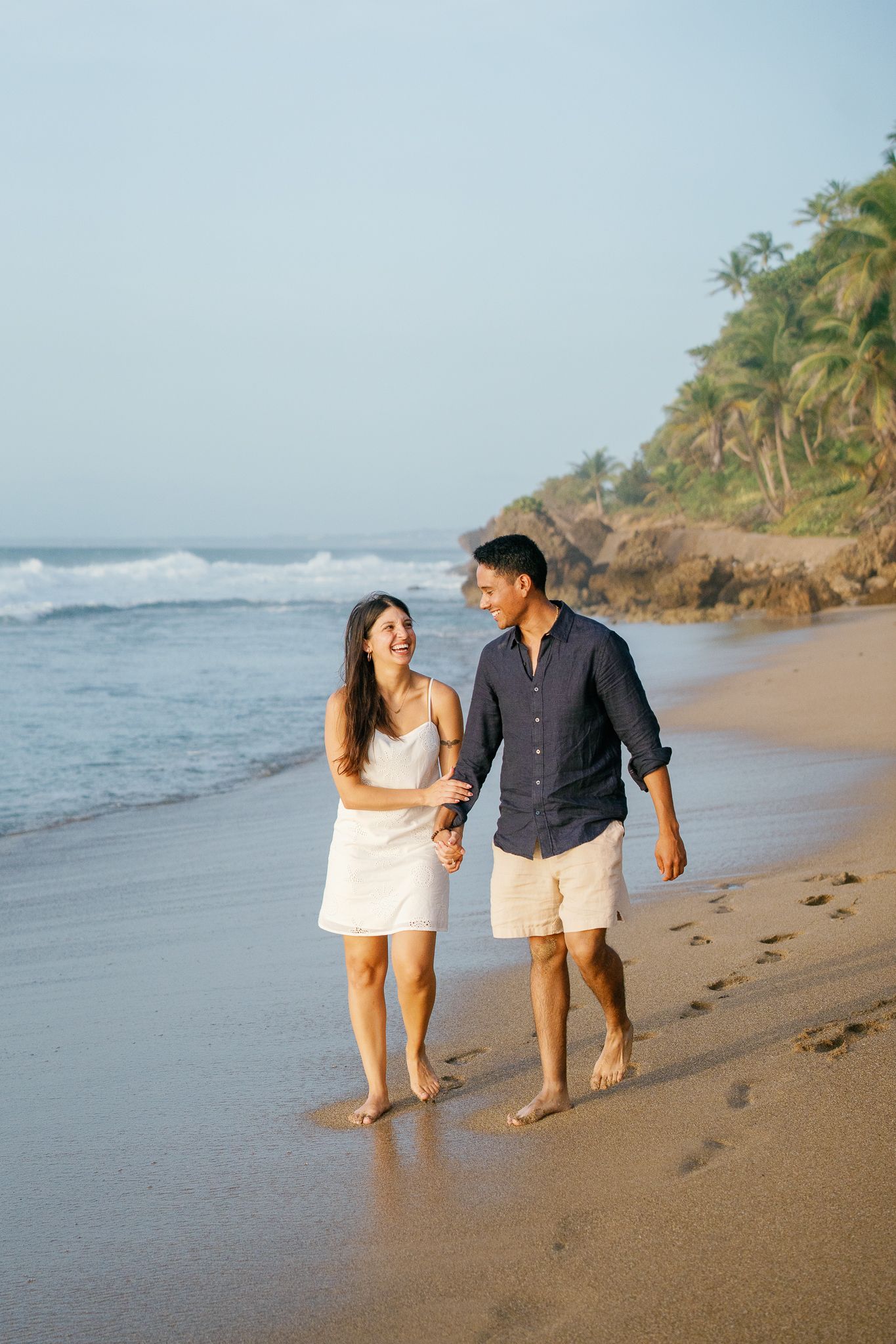 A couple walks together on a tropical beach with palm trees and ocean waves in the background.
